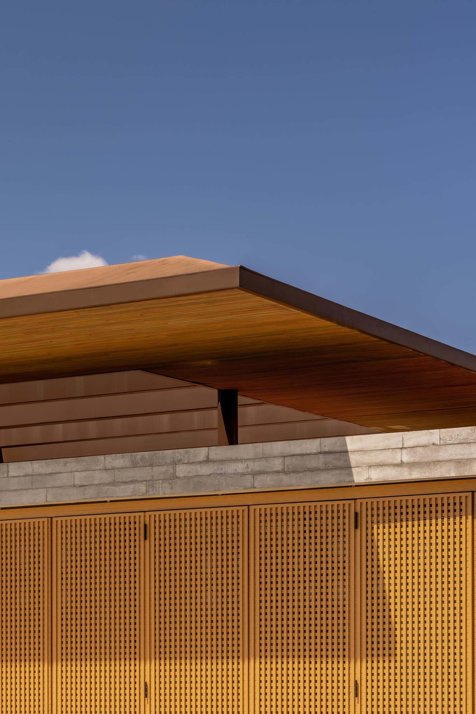 Close-up view of a modern building facade featuring warm wooden perforated screens, a thick concrete eave, and horizontal wood cladding against a clear blue sky.