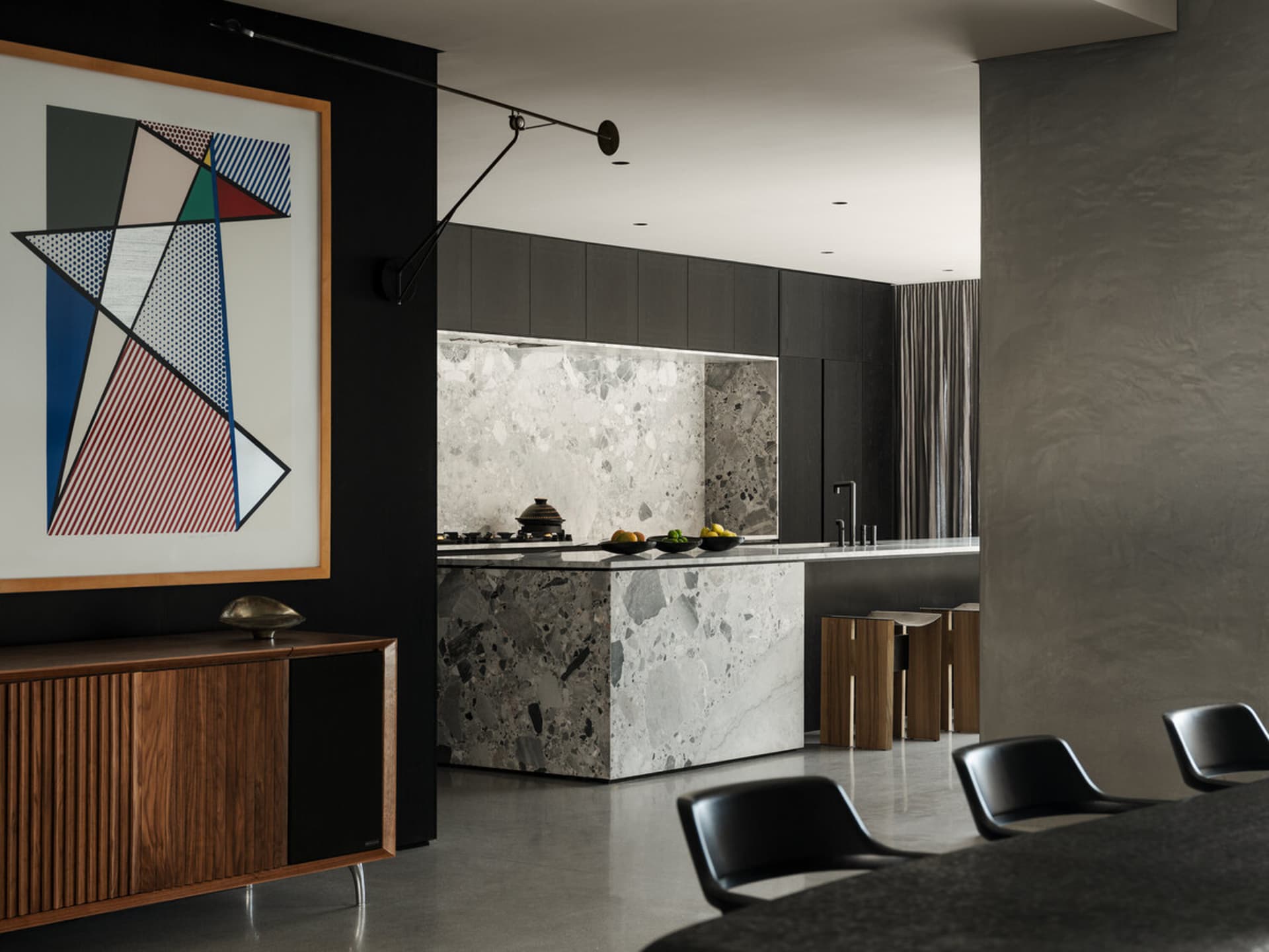 Modern dark kitchen interior with large white and grey terrazzo island, dark wood cabinetry, black dining chairs, and a large colorful geometric abstract painting on a black wall.