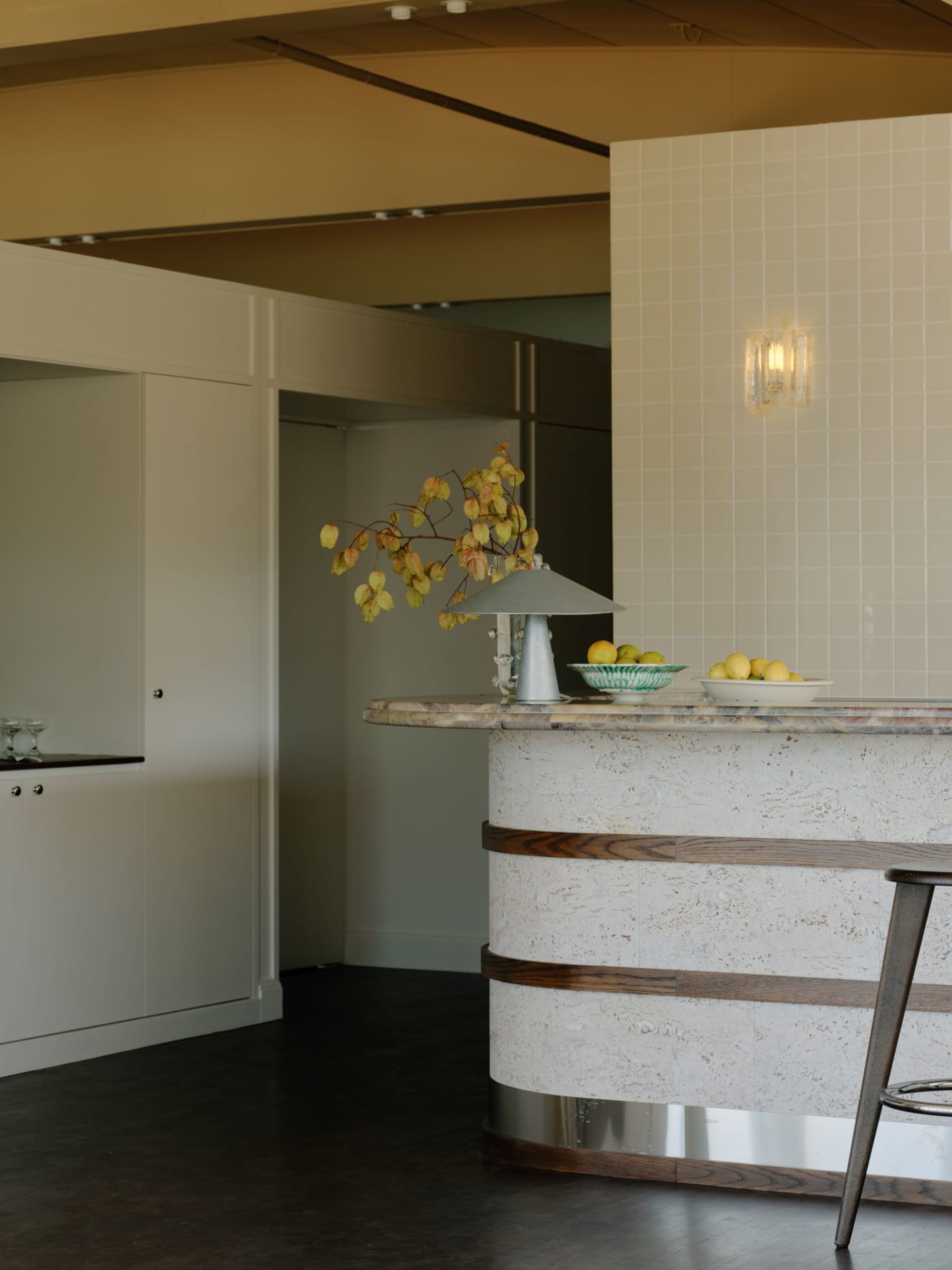 Modern kitchen with a curved travertine island featuring horizontal oak bands, a grey table lamp, fruit bowls, and a wall sconce against a white tiled wall.