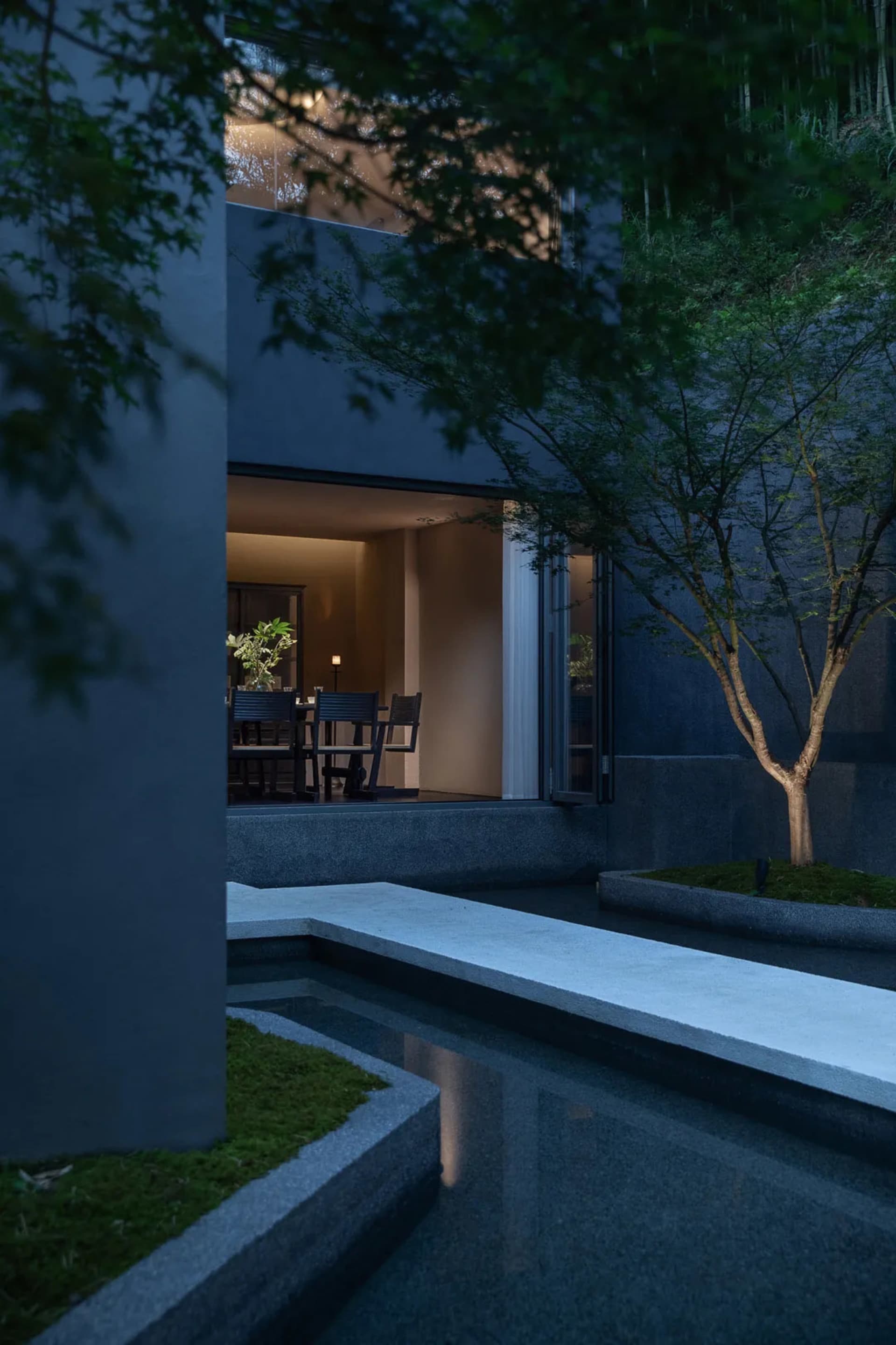 Night view of a modern concrete house courtyard with a linear water feature, moss garden, small tree, and warmly lit indoor dining area visible through large openings.