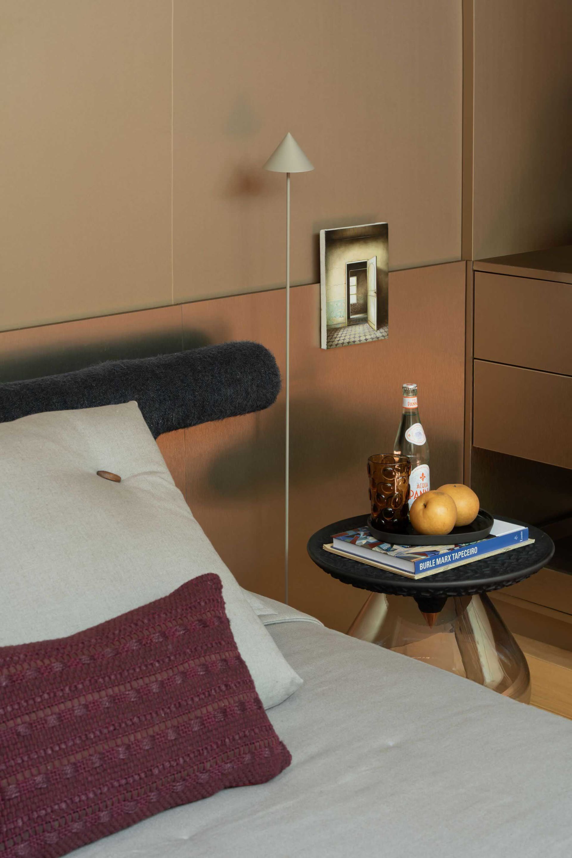 Modern bedroom corner featuring a bed with white and burgundy pillows, a black textured side table with brass base, a minimalist white floor lamp, and warm metallic wall panels.