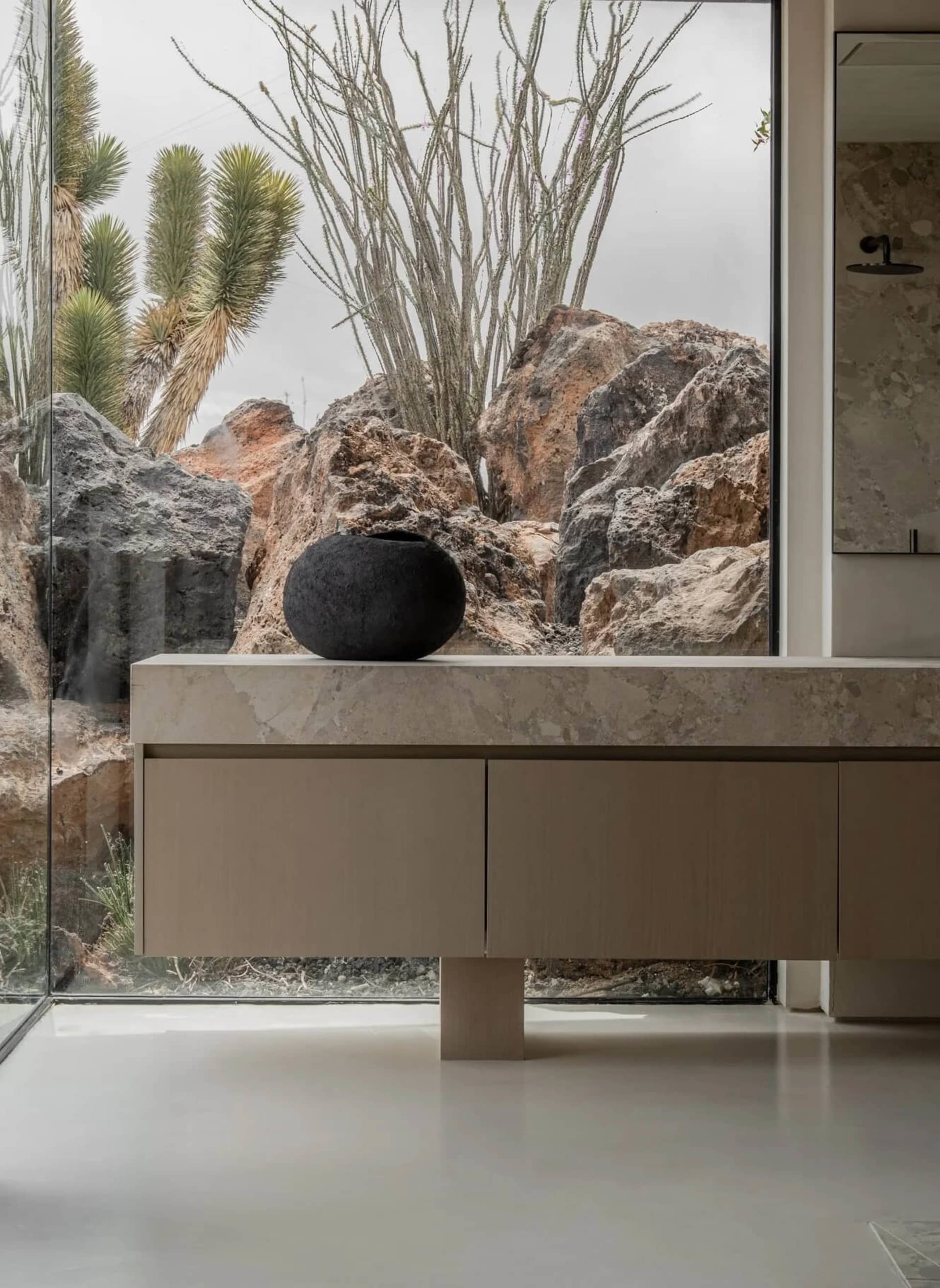 Modern minimalist bathroom featuring a floating travertine vanity with a black ceramic vessel, large glass window overlooking dramatic desert rocks and cacti.