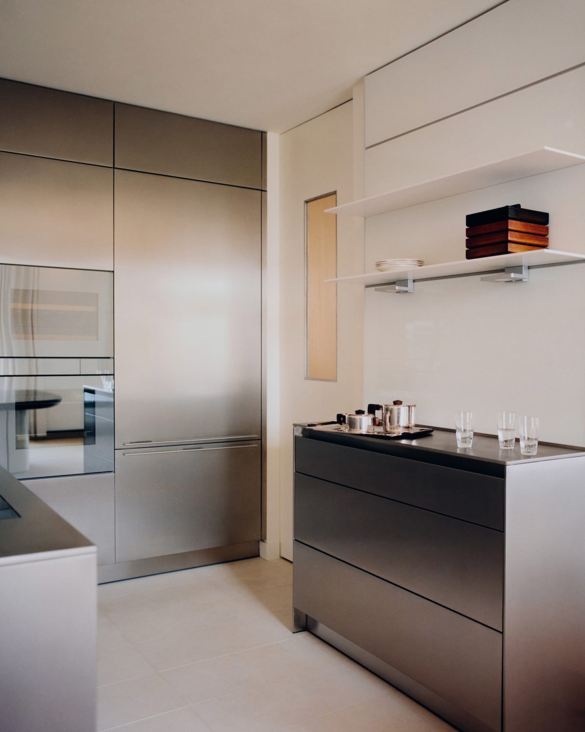 Modern minimalist kitchen featuring seamless stainless steel cabinetry, built-in appliances, dark stone countertop, and white floating shelves with wooden storage boxes.