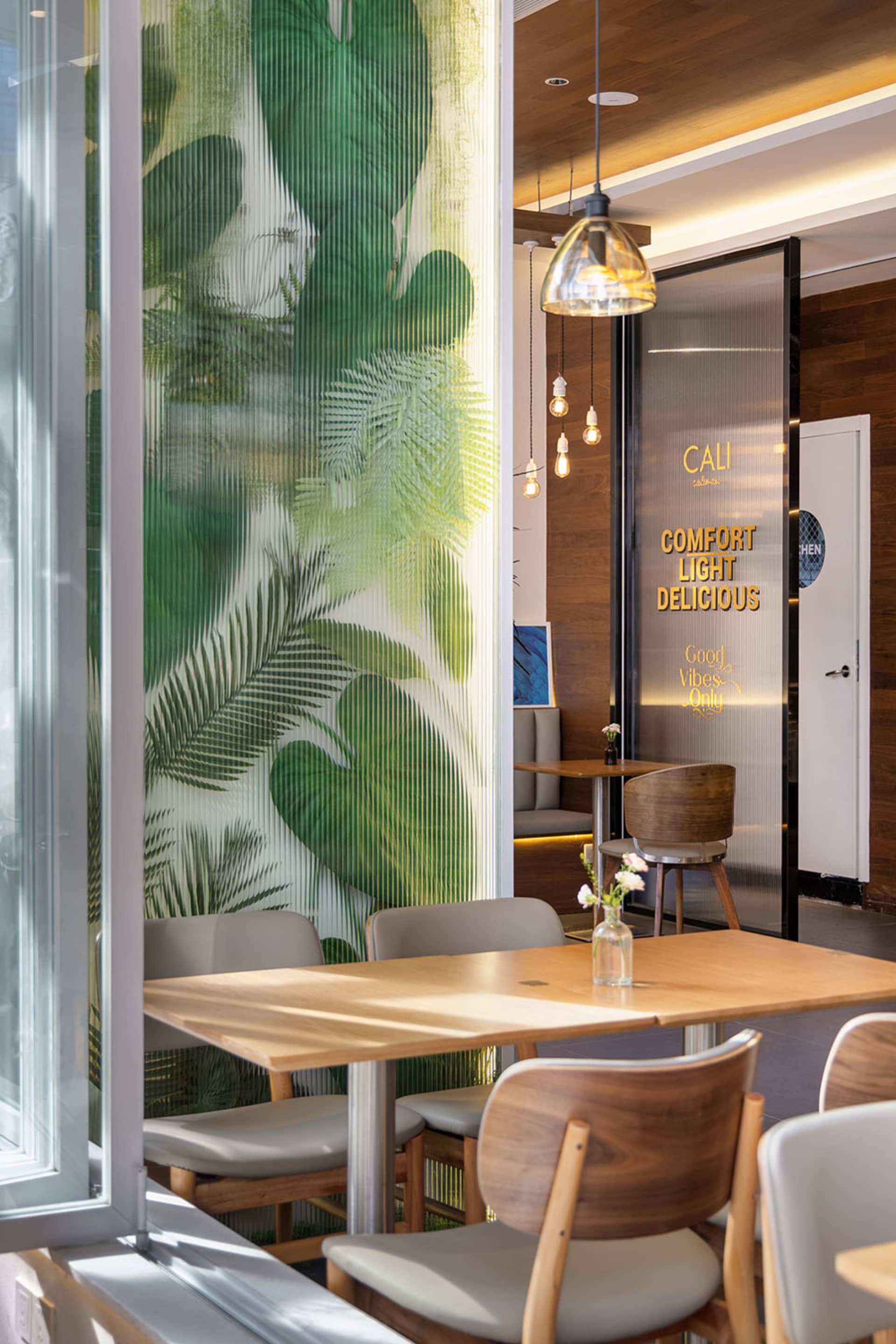 Modern restaurant dining area featuring a large tropical leaf mural on a ribbed glass partition, light wood tables, and mixed wood and upholstered chairs under pendant lighting.