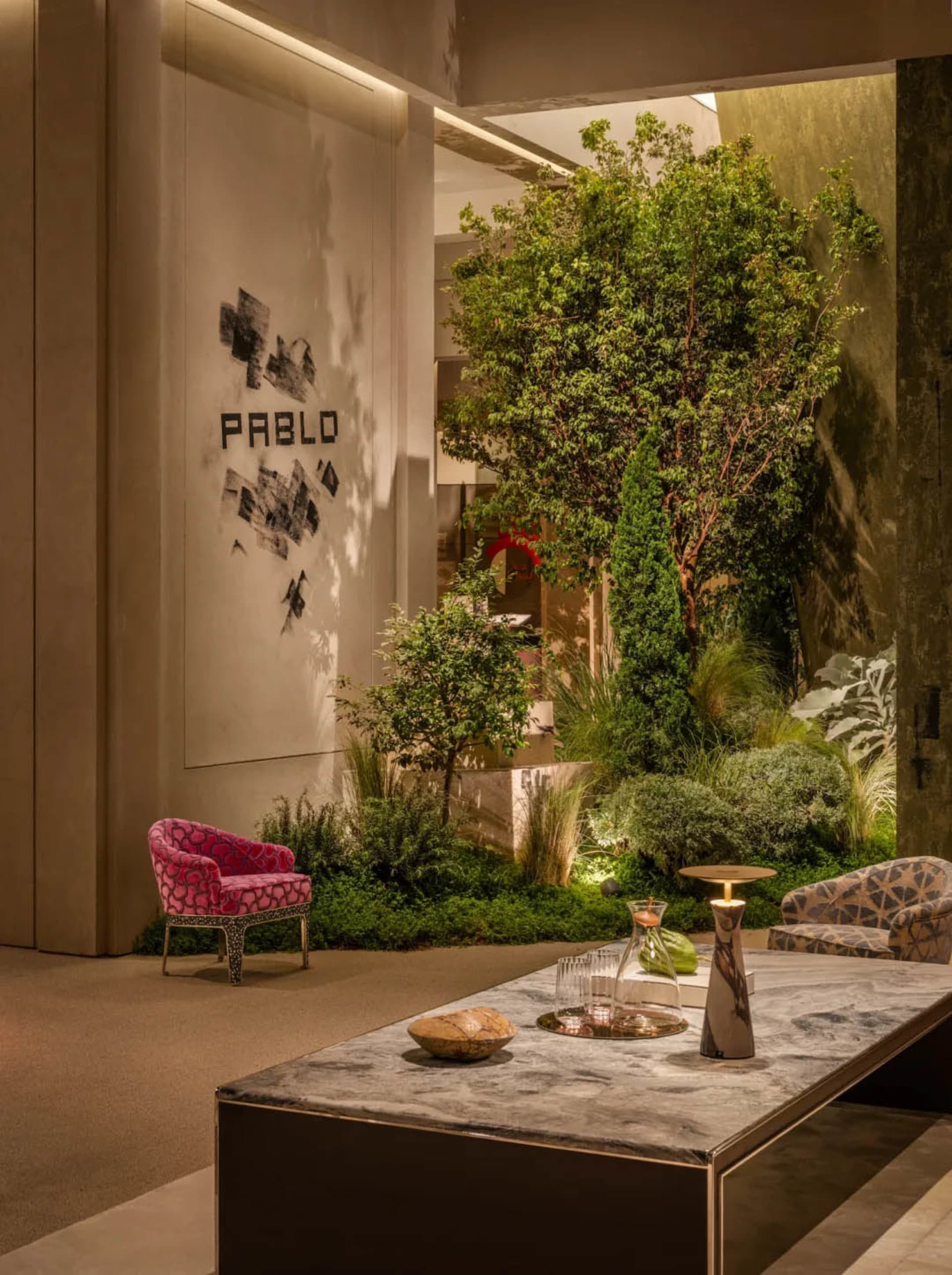 Modern lobby lounge featuring a large indoor garden with lush trees and plants, a marble coffee table, a vibrant pink patterned armchair, and a geometric patterned sofa under warm ambient lighting.