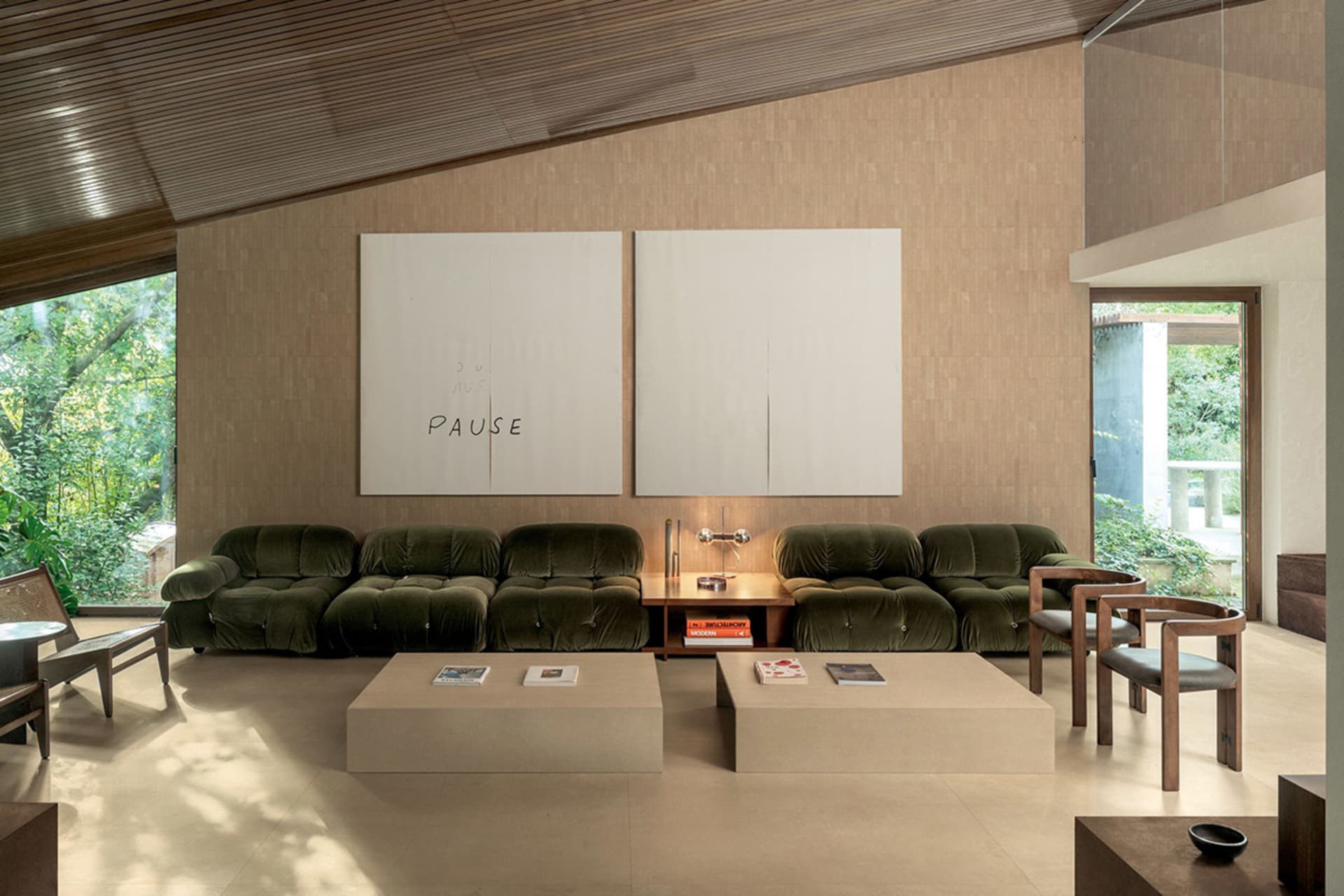 Modern living room featuring two forest green velvet modular sofas, beige stone coffee tables, warm wood panel walls, large abstract white artwork with the word PAUSE, and views of lush greenery outside.