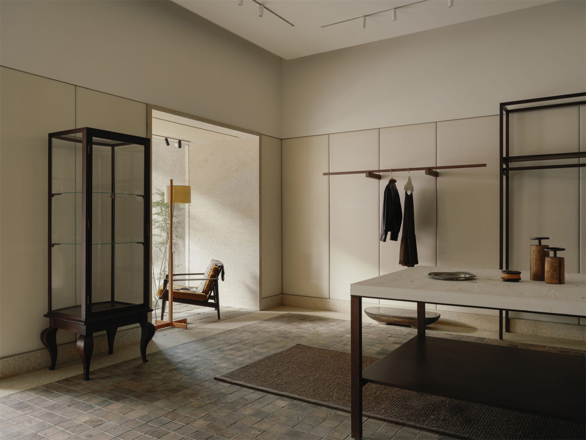 Modern minimalist entryway featuring a tall dark walnut glass vitrine, a limestone-top console table, open wardrobe system, and a vintage armchair visible in the adjacent sunlit room.