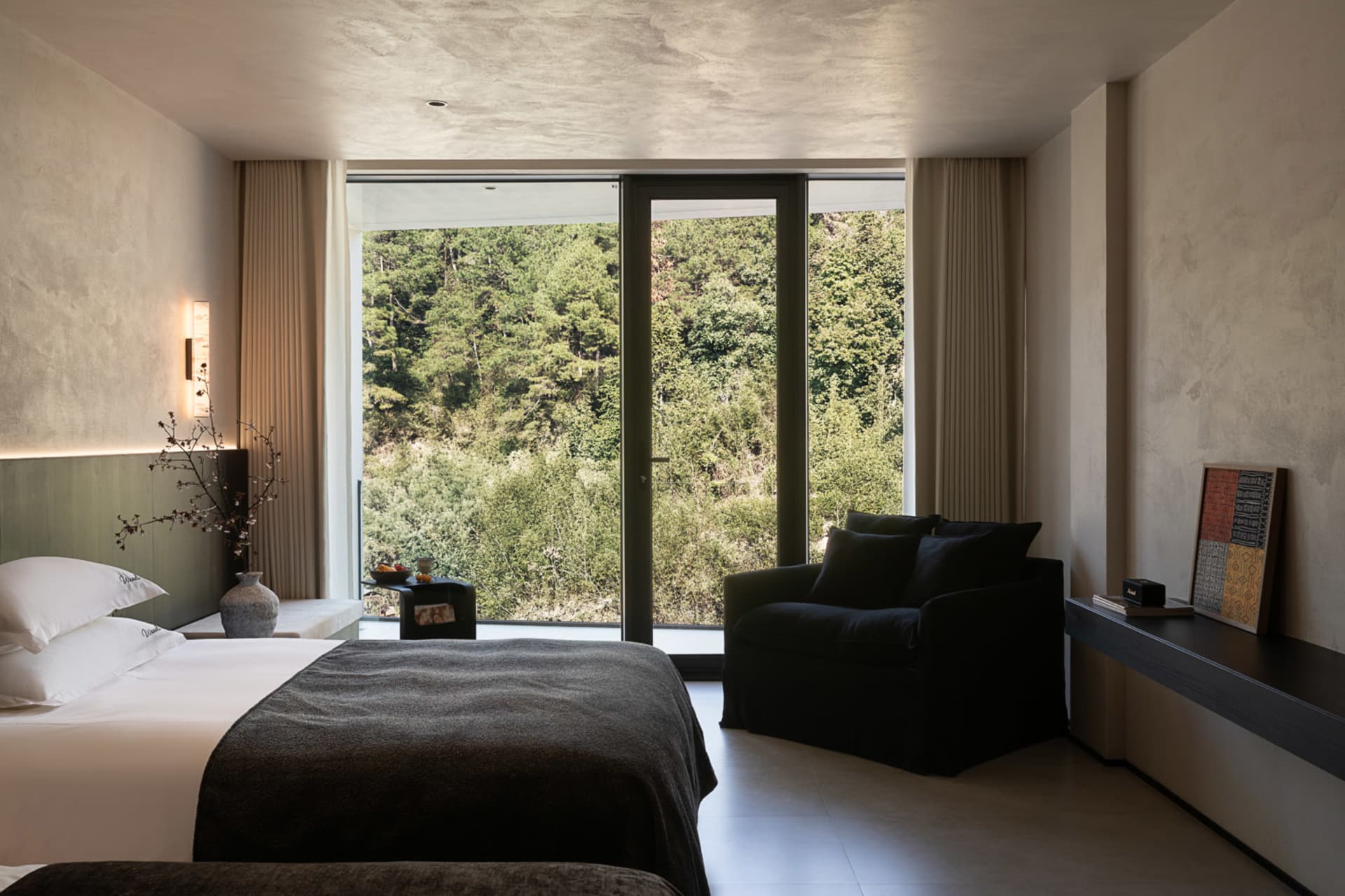 Modern minimalist bedroom featuring a large bed with white linens and dark throw, black armchair, textured plaster walls, and expansive glass doors opening to a dense forest view.