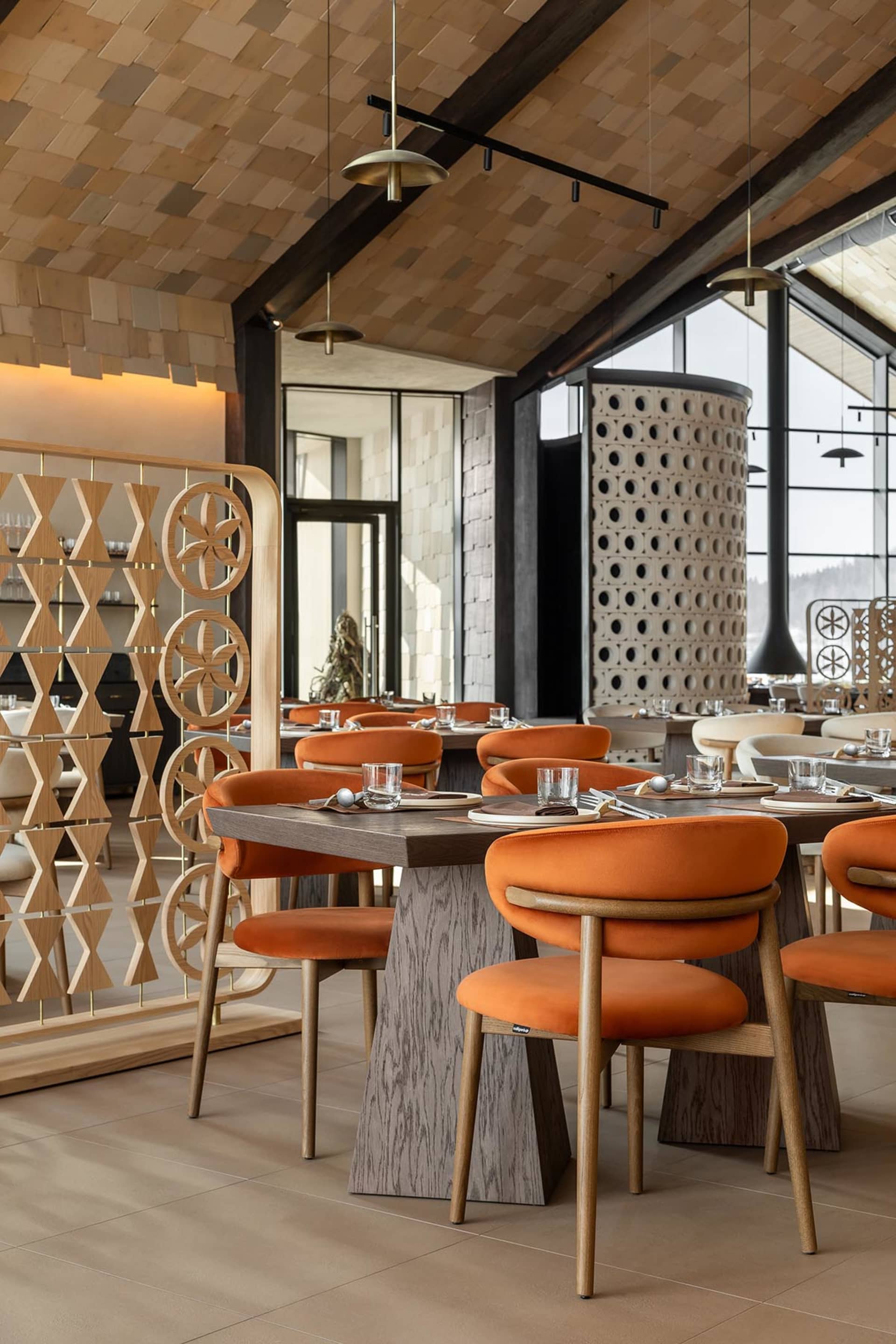 Modern restaurant interior with burnt orange velvet dining chairs, dark wood tables, geometric wooden room dividers, and a striking wood-paneled ceiling with pendant lights.
