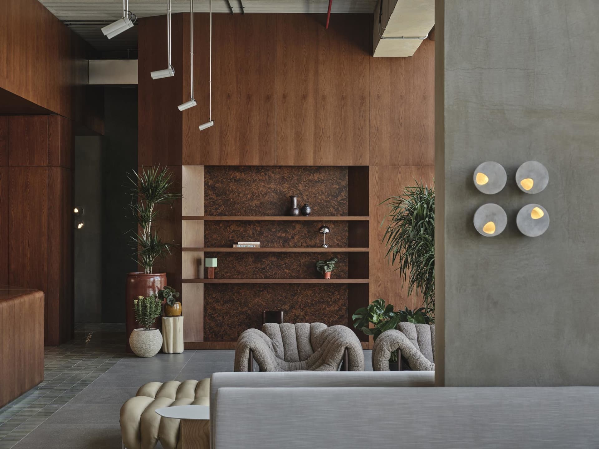 Modern lobby interior featuring a large walnut burl wood feature wall, built-in shelving, two curved bouclé armchairs, concrete wall sconces, and multiple potted plants.