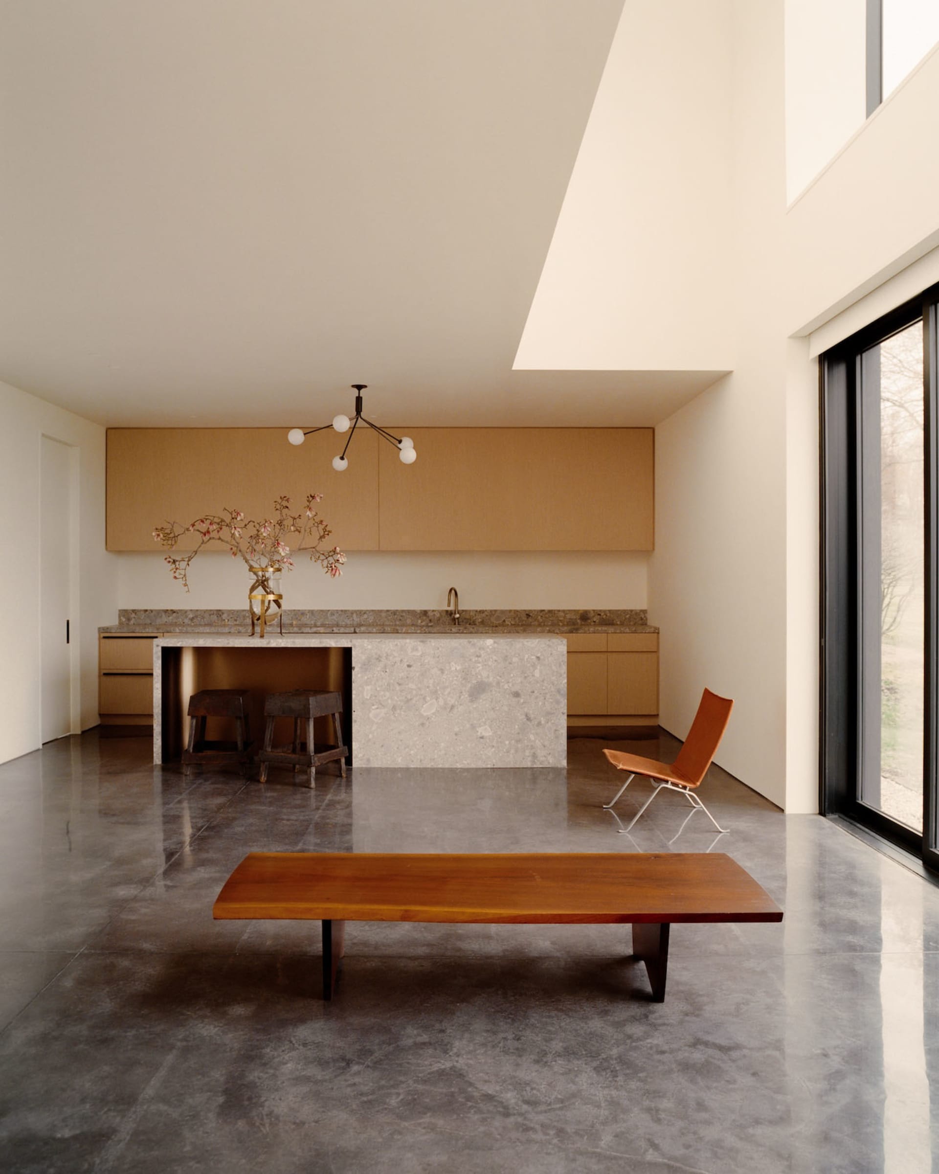 Modern minimalist open-plan kitchen and living area featuring polished concrete floors, a terrazzo kitchen island, light oak cabinetry, a live-edge oak bench, and a sculptural black chandelier.