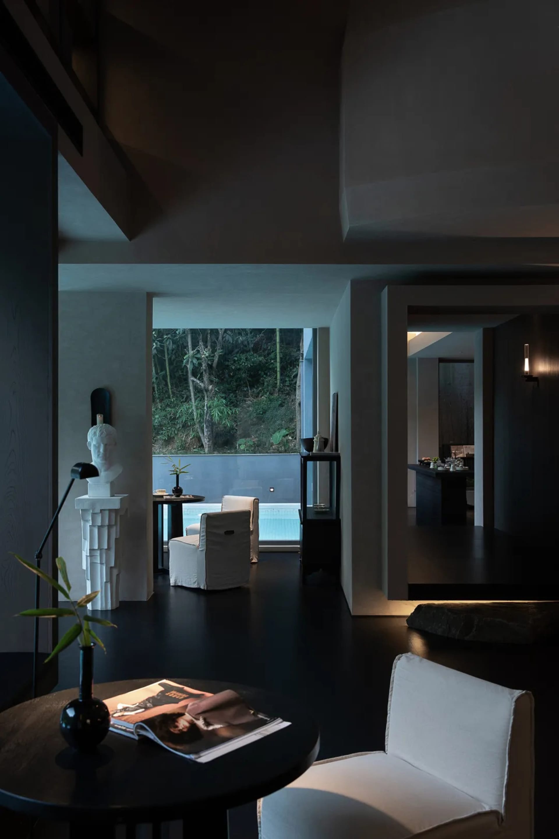 Modern minimalist living room featuring dark concrete walls, black floor, white linen armchairs, sculptural white bust on pedestal, and large opening revealing a swimming pool surrounded by lush greenery.