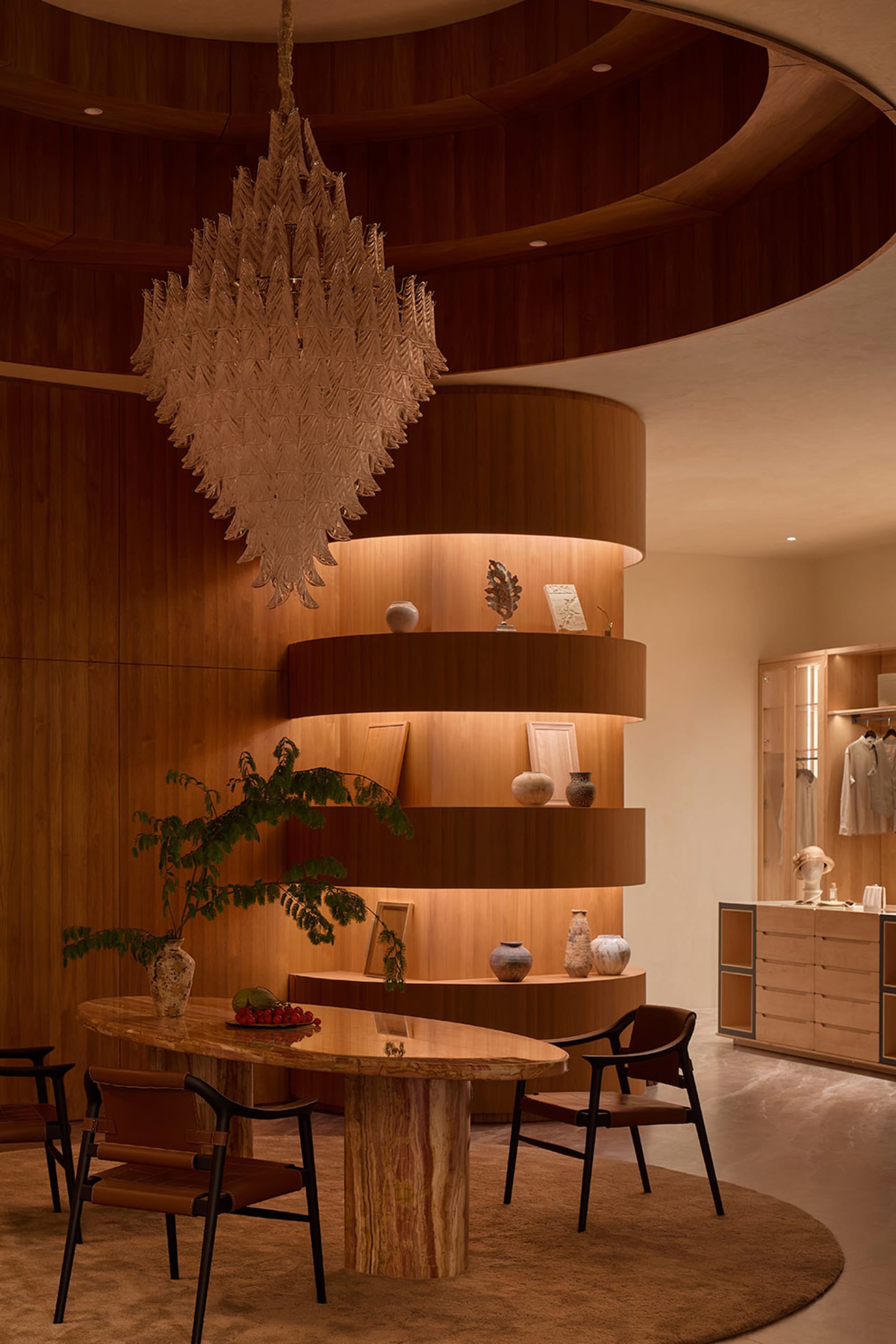 Modern luxury dining room featuring a large oval onyx marble table, brown leather dining chairs, a massive sculptural chandelier, and a glowing curved multi-level wooden display wall with decorative objects.