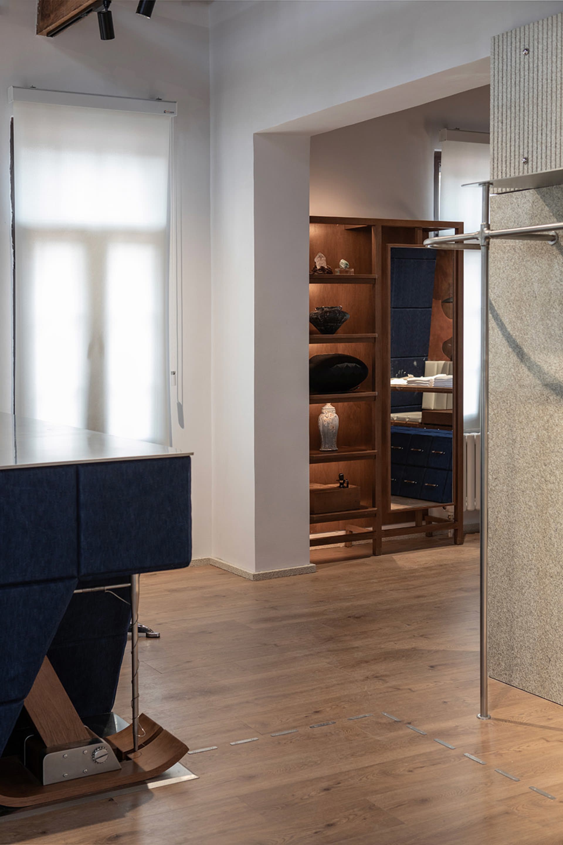 Modern minimalist entryway featuring a blue upholstered bench with walnut base, built-in walnut display shelves with decorative objects, light wood flooring, and a textured beige wall panel.