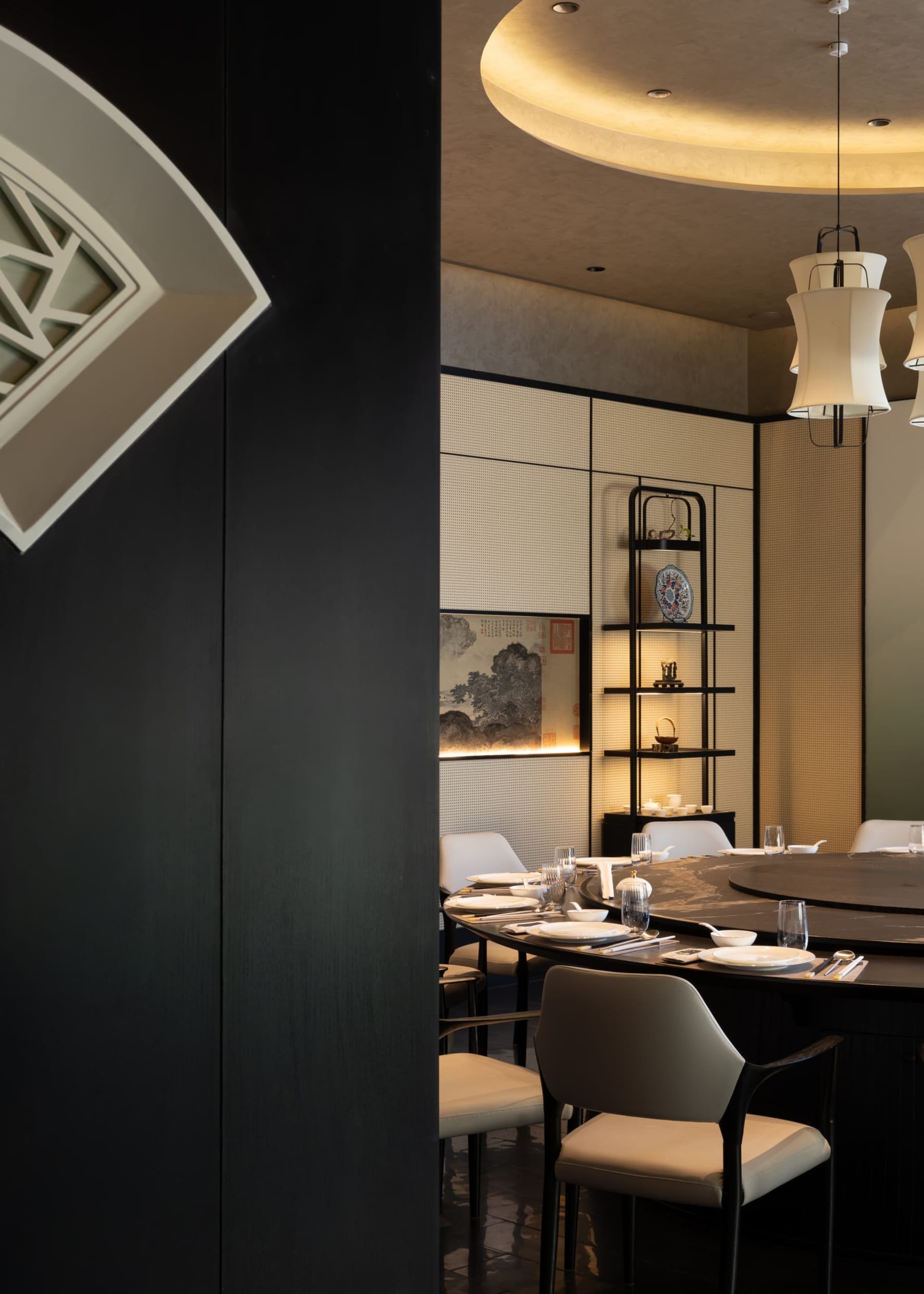 Elegant Chinese restaurant interior with a large round dark dining table, cream upholstered chairs, silk lanterns, and traditional Chinese artwork on a textured wall.