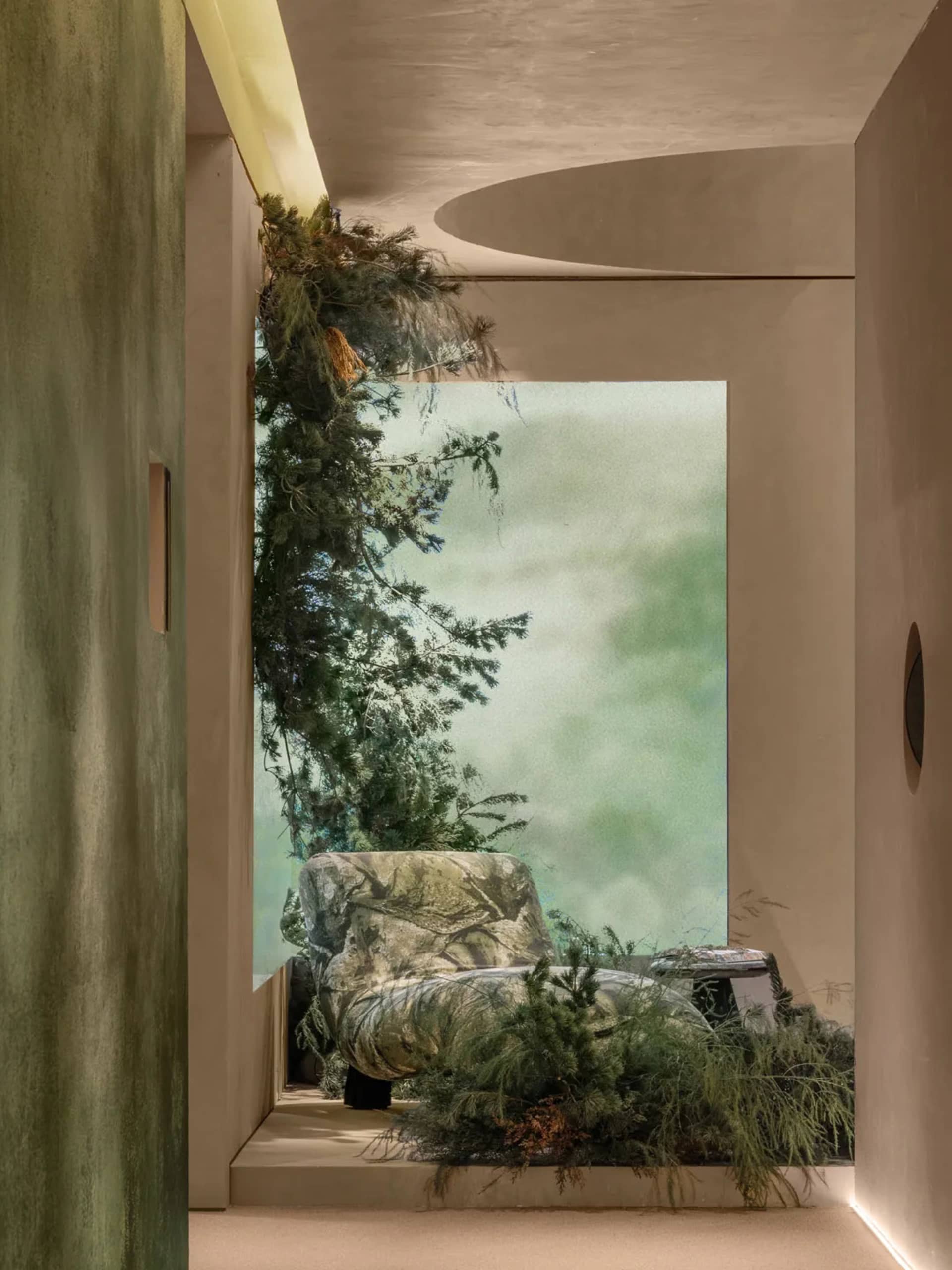 Modern biophilic interior featuring a stone-like armchair surrounded by lush evergreen plants, with soft natural light filtering through a large opening and curved plaster architecture.