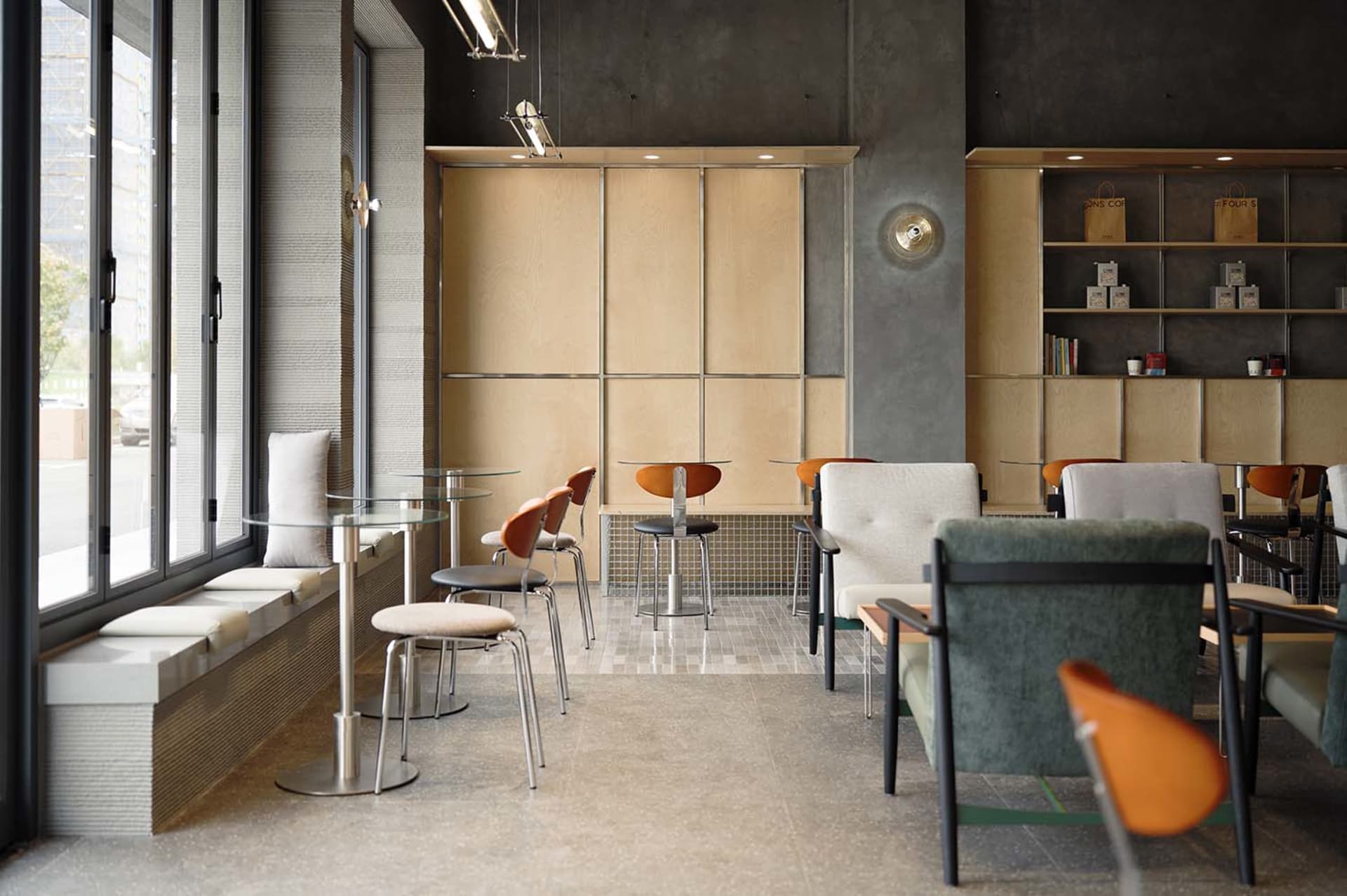 Modern cafe interior featuring concrete walls, light wood cabinetry, built-in window seating, round glass tables, and a mix of upholstered and molded wood chairs in green, orange, and neutral tones.