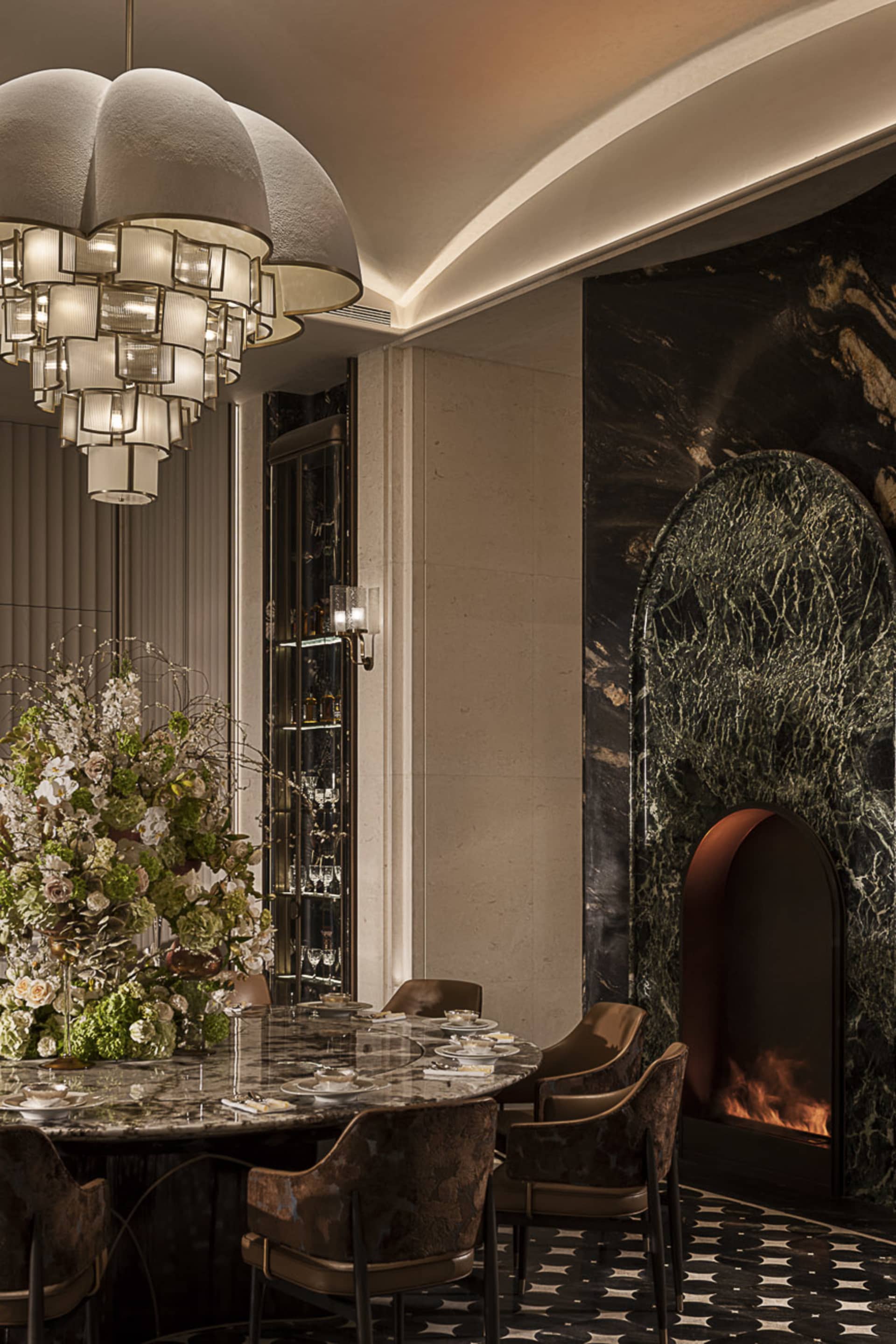 Luxurious dining room featuring a large round marble table, velvet patterned dining chairs, a striking green marble fireplace with arched opening, and an oversized cloud-like chandelier with multiple lit tiers.