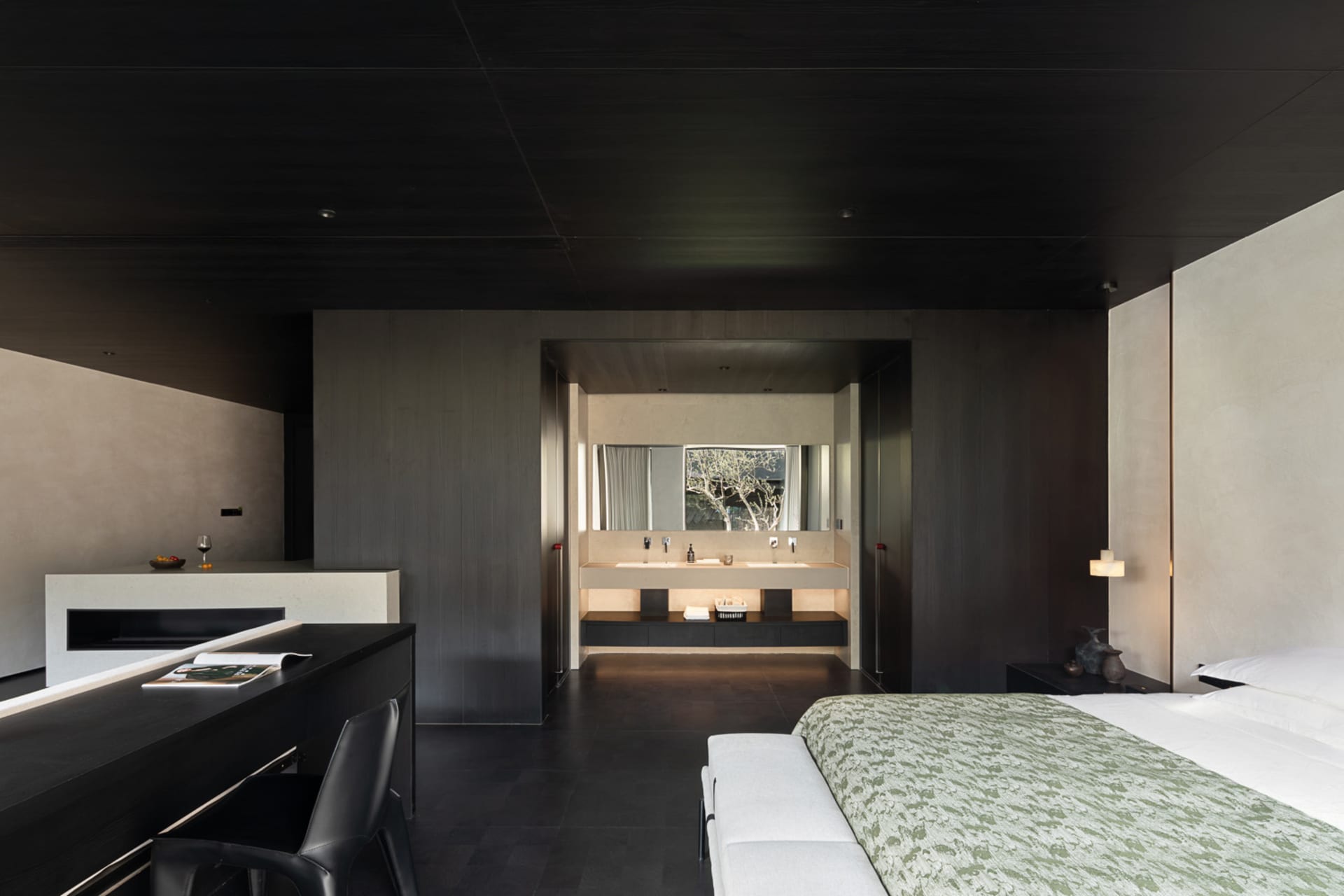 Modern dark-themed bedroom featuring a large bed with green patterned coverlet, black desk and chair in the foreground, and an open en-suite bathroom with double vanity visible in the background.