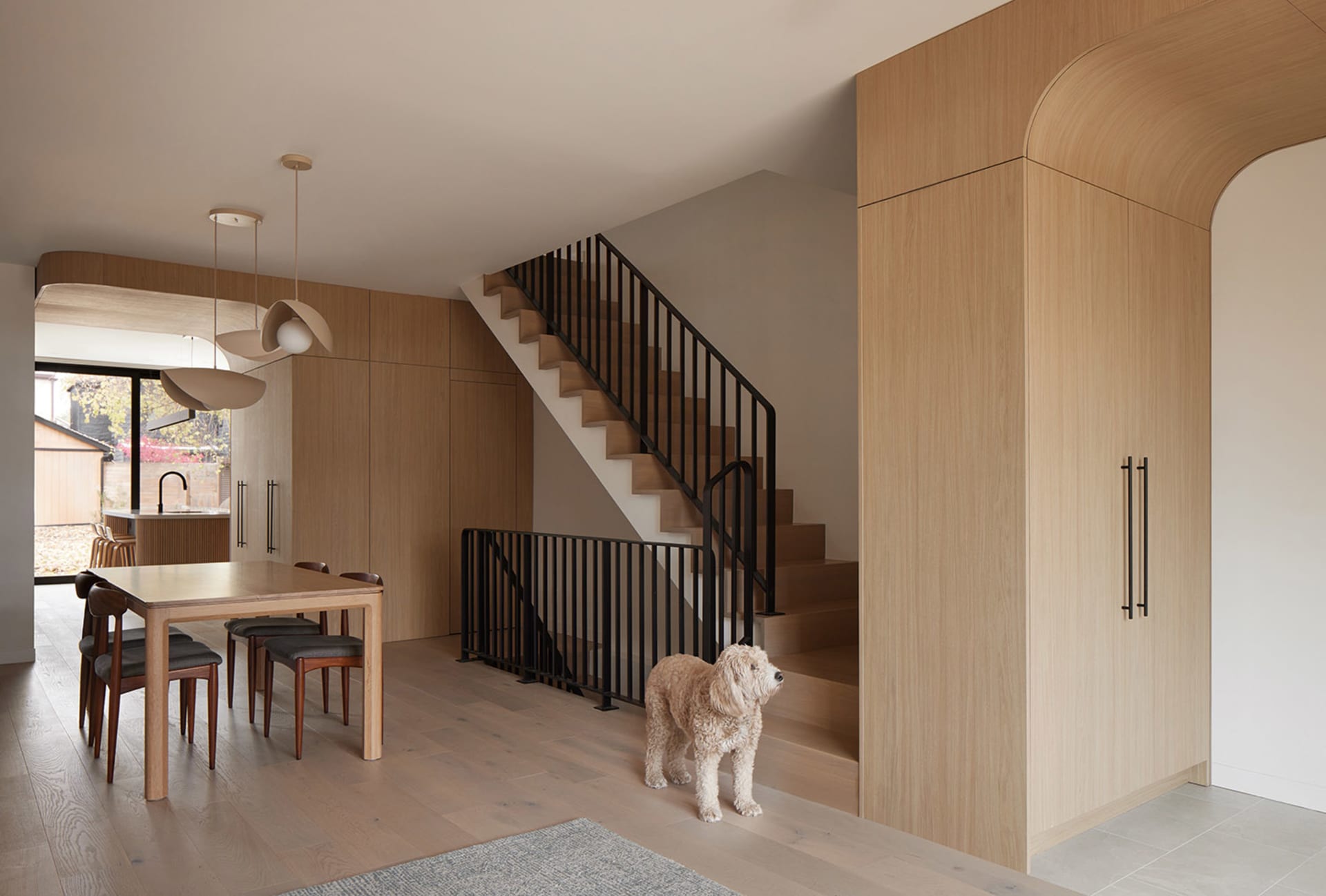 Modern dining room featuring a solid oak table and chairs, three pendant lights, light oak wall paneling, a black metal railing staircase, and a curly-haired dog standing on light wood flooring.