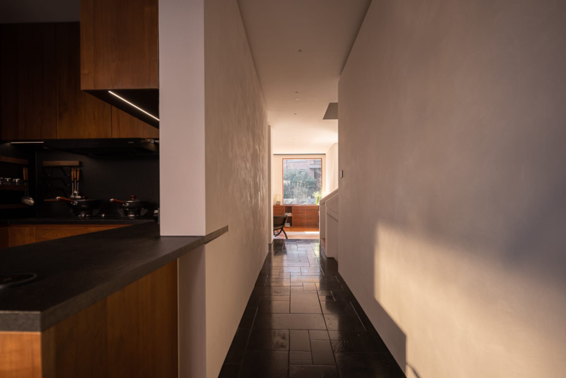 View from a modern walnut kitchen counter looking down a narrow hallway with dark tiled floor toward a bright window at the far end.