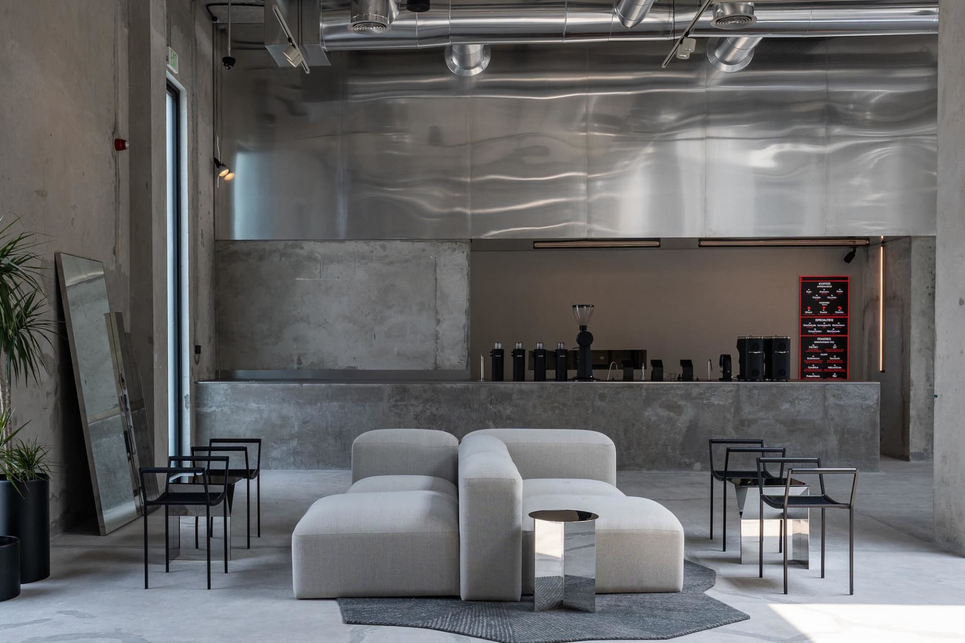 Modern industrial cafe lounge with raw concrete walls, a large rippled stainless steel feature wall, light grey modular sofas, black metal chairs, and exposed ceiling ducts.