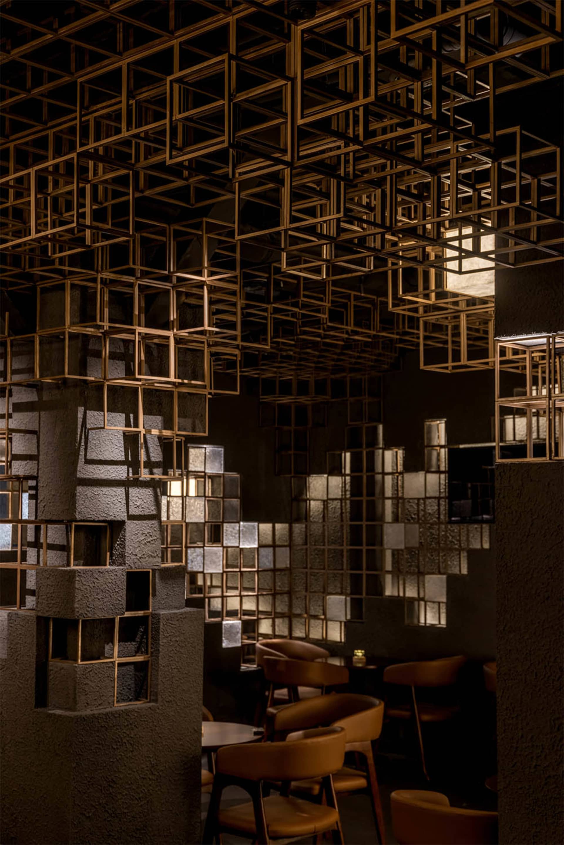 Interior of a modern restaurant featuring a dramatic ceiling installation of interlocking brass cuboid frames, textured concrete pillars with integrated lighting, and warm brown leather dining chairs.