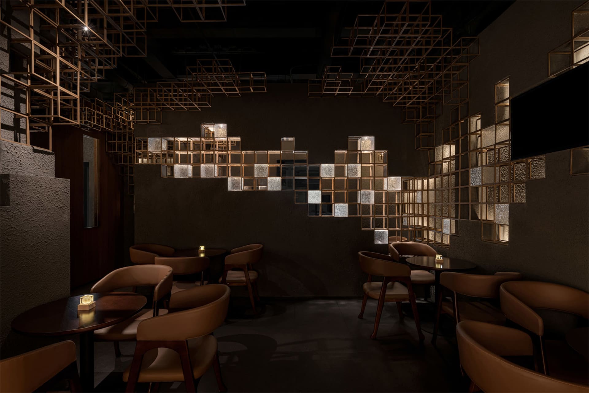 Dimly lit modern restaurant interior featuring brown leather dining chairs, dark round tables, and a striking illuminated brass cube grid art installation covering the back wall.