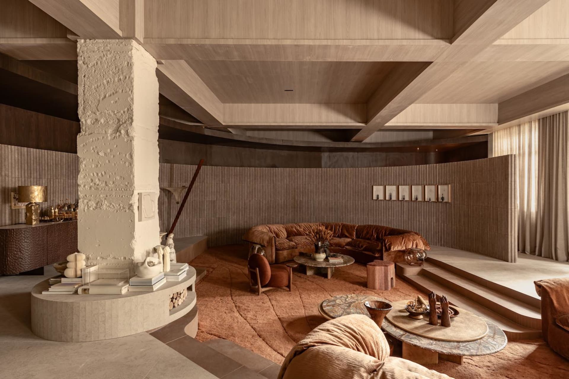 Modern living room featuring a large curved terracotta velvet sofa, a prominent white textured plaster column, layered circular coffee tables, and warm earthy tones throughout the space.