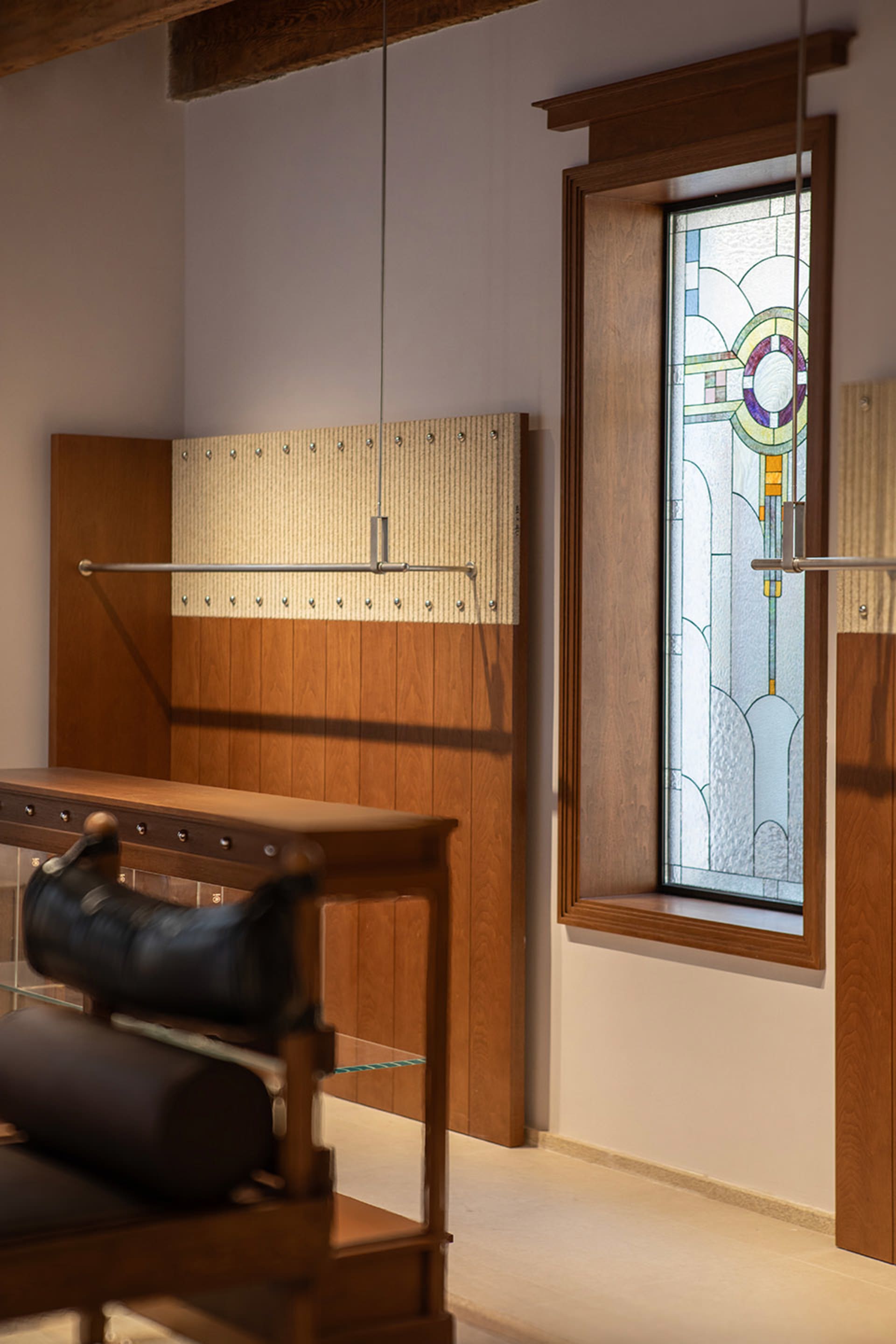 Walnut wood hall tree with metal hanging rod and textured panel, positioned next to a tall wooden-framed stained glass window featuring Art Nouveau patterns in purple, green and yellow.