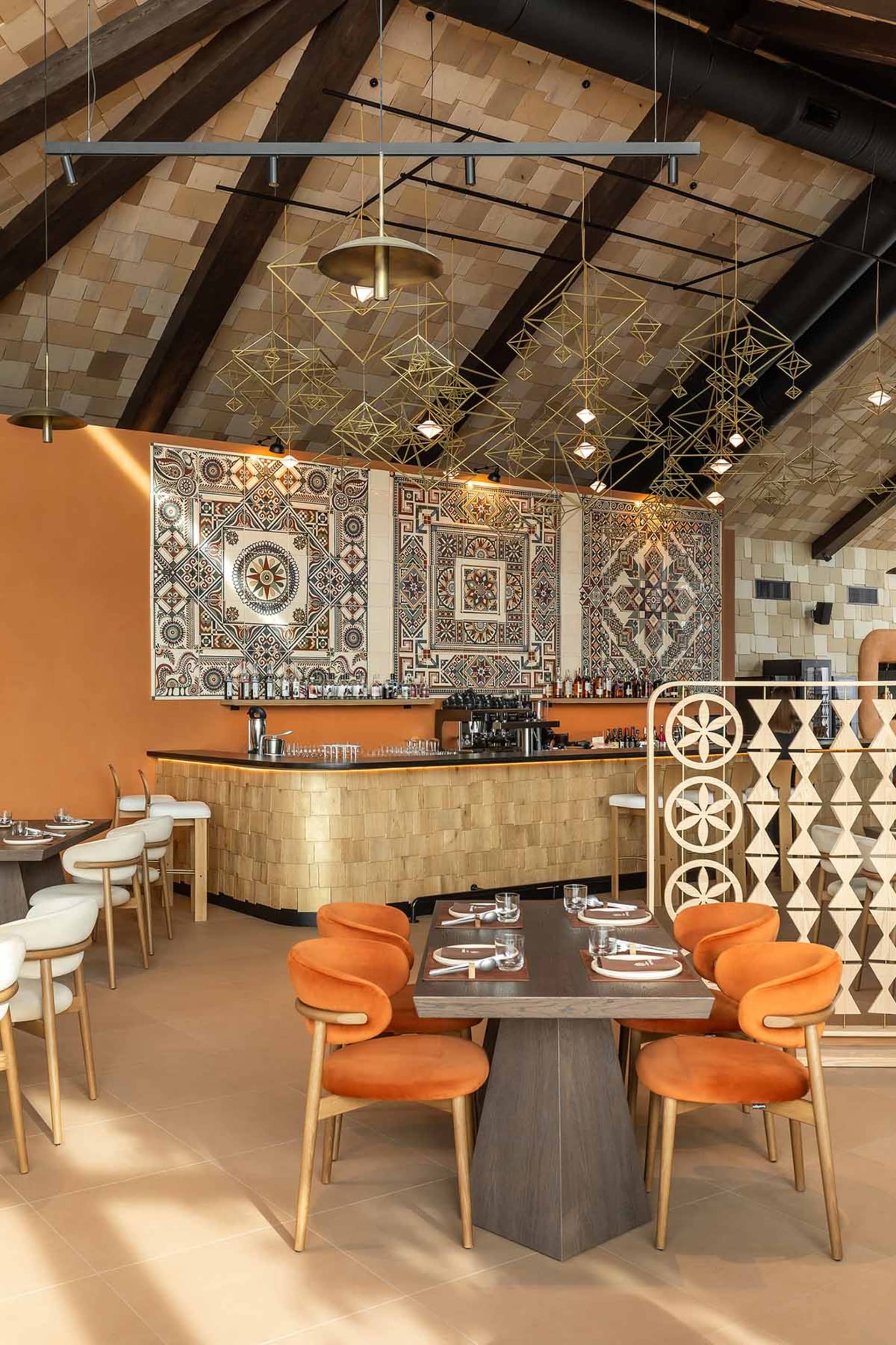 Modern restaurant interior with orange velvet dining chairs, dark wood tables, a curved bar with golden tile cladding, large colorful patterned tiles on the wall, and geometric brass pendant lights hanging from a high beamed ceiling.