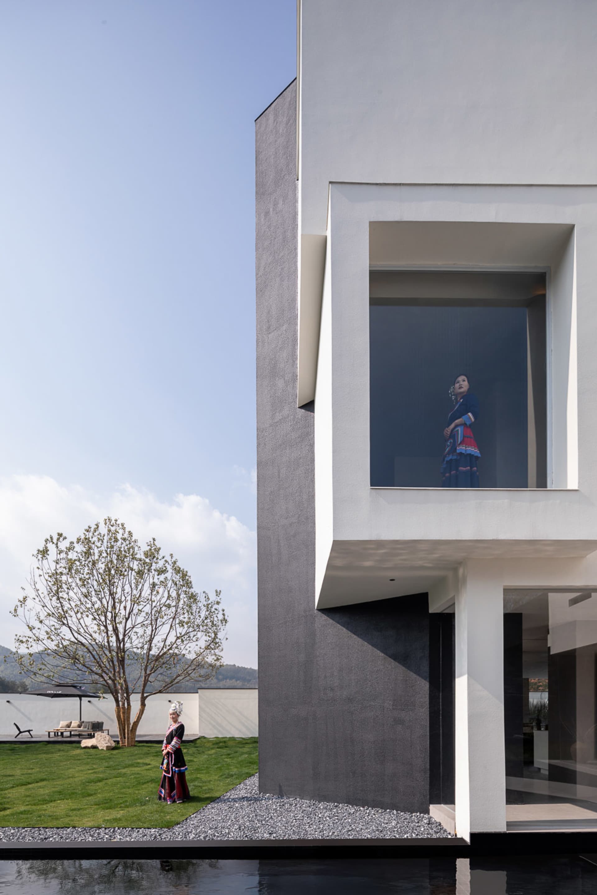 Modern minimalist concrete villa exterior with large square window framing a woman in colorful traditional indigenous clothing, a second woman in similar attire standing on the lawn beside a young tree, with a reflecting pool in the foreground.
