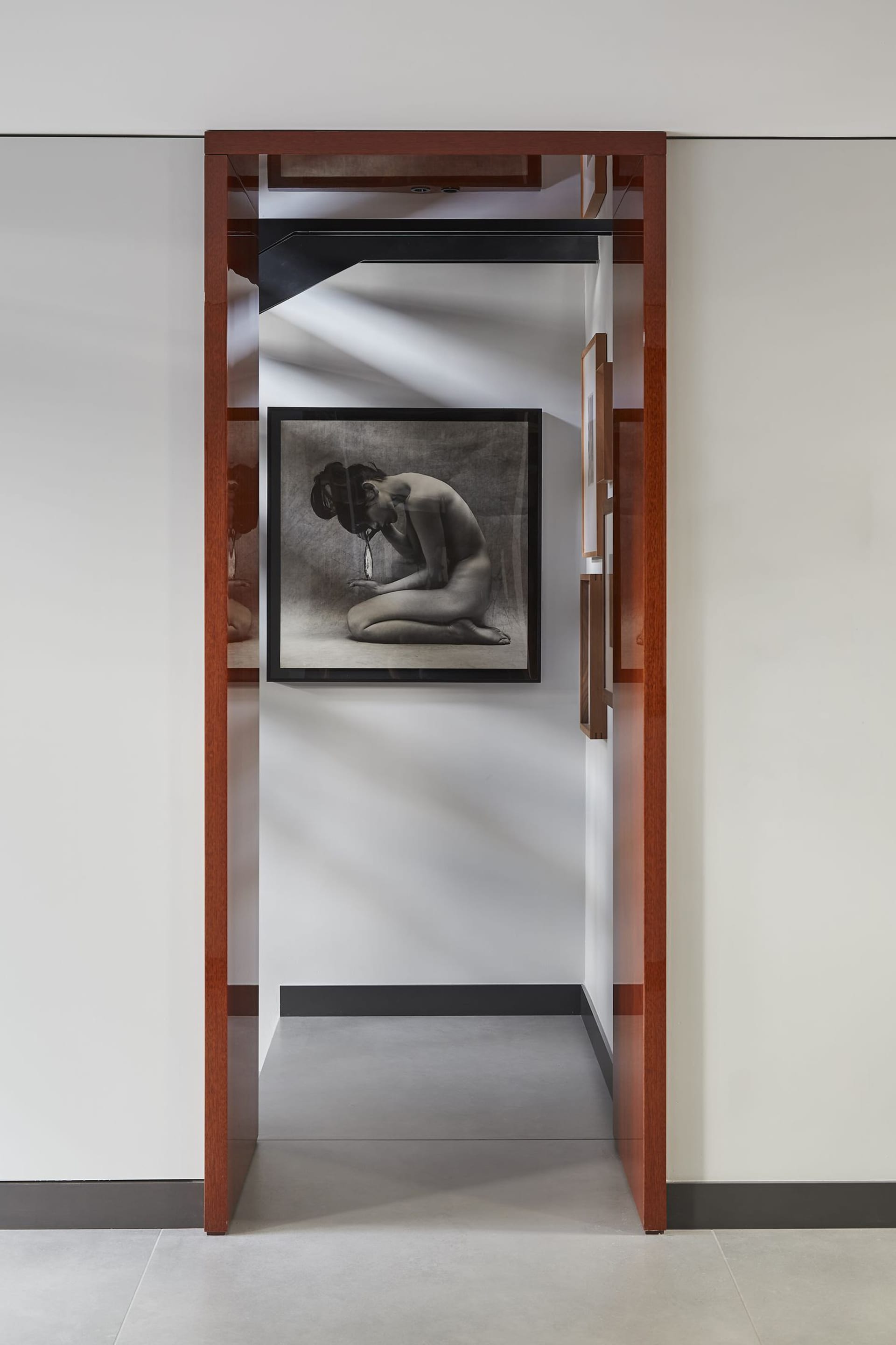 Modern minimalist hallway featuring a large framed black and white nude photograph, rich walnut wood door frames, and dramatic diagonal light patterns on white walls.