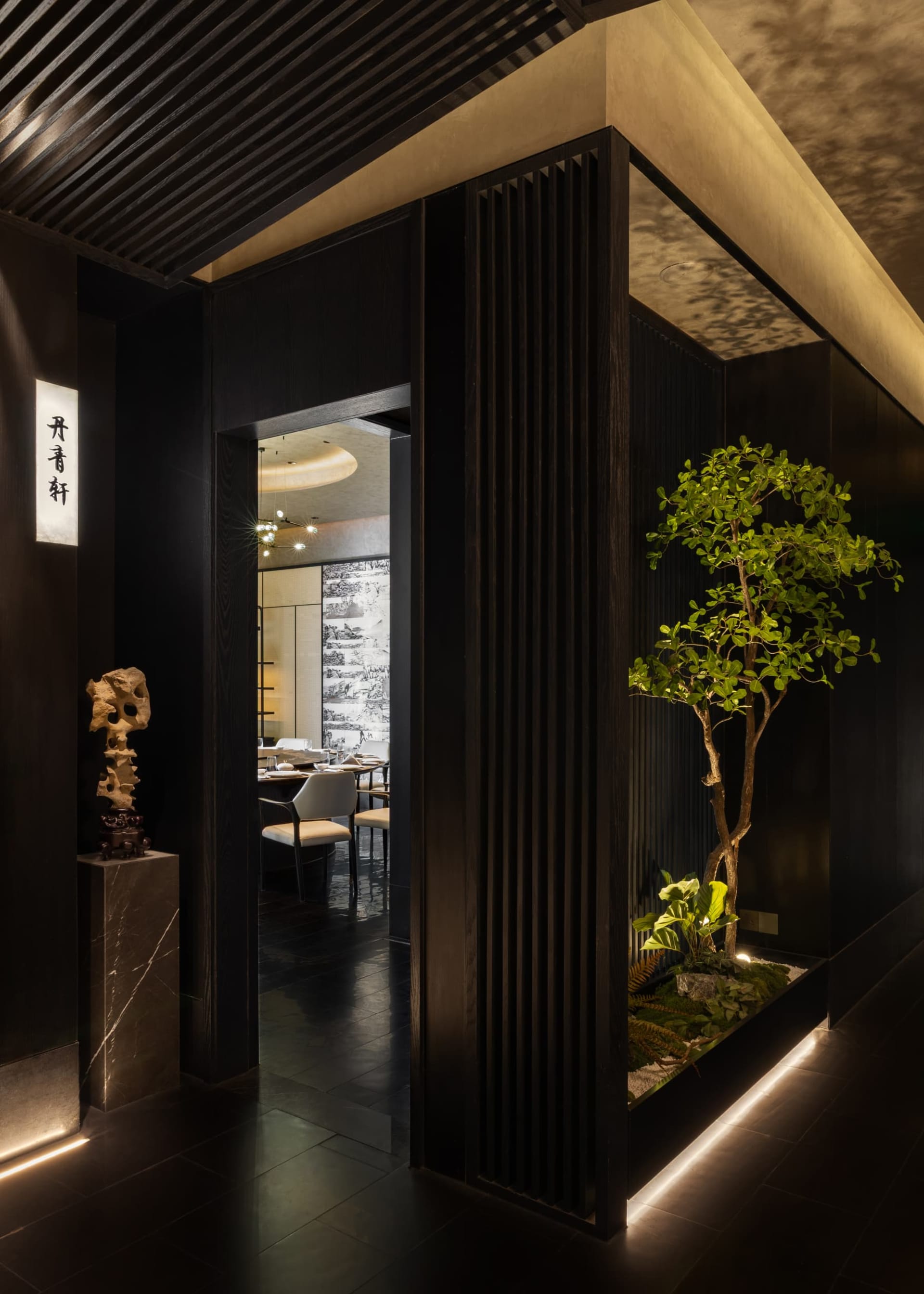 Modern restaurant entrance featuring vertical dark wood slats, an indoor tree garden with uplighting, a scholar rock sculpture on a marble pedestal, and a view into the dining area.