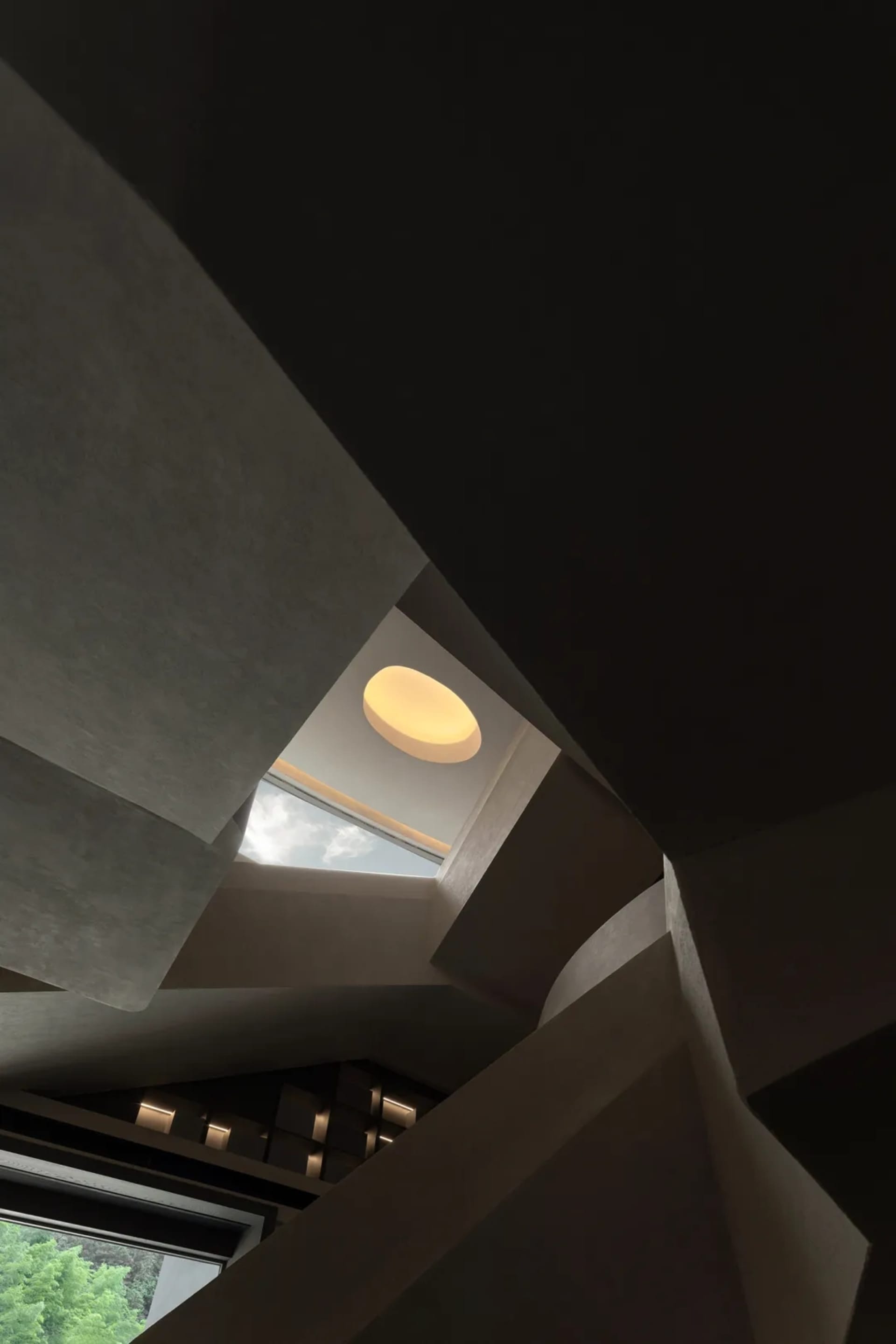 Low-angle view looking up at a sculptural concrete staircase featuring sharp geometric angles, a large circular skylight, and warm recessed lighting.