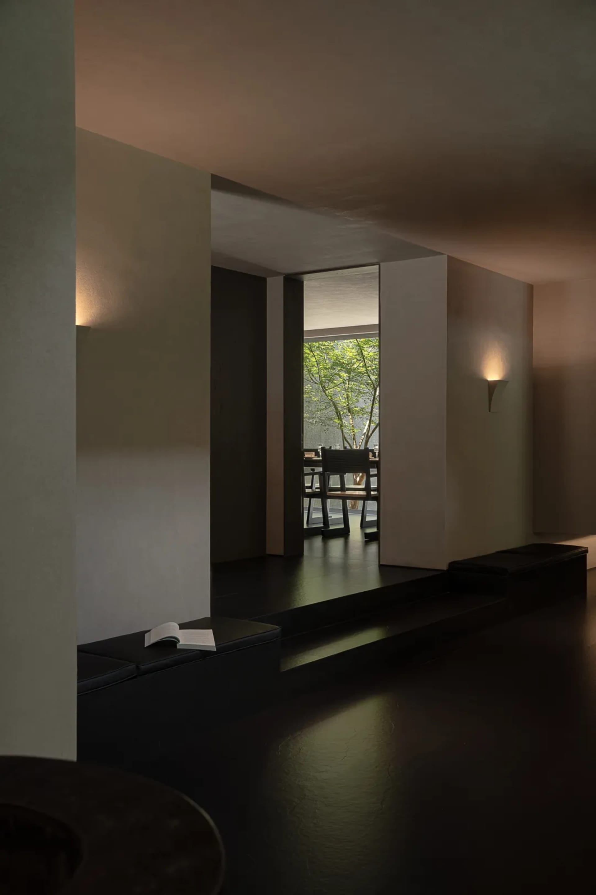 Modern minimalist interior featuring a dark concrete reading nook with built-in black leather bench, open book, and view to a lush green garden through a large opening.