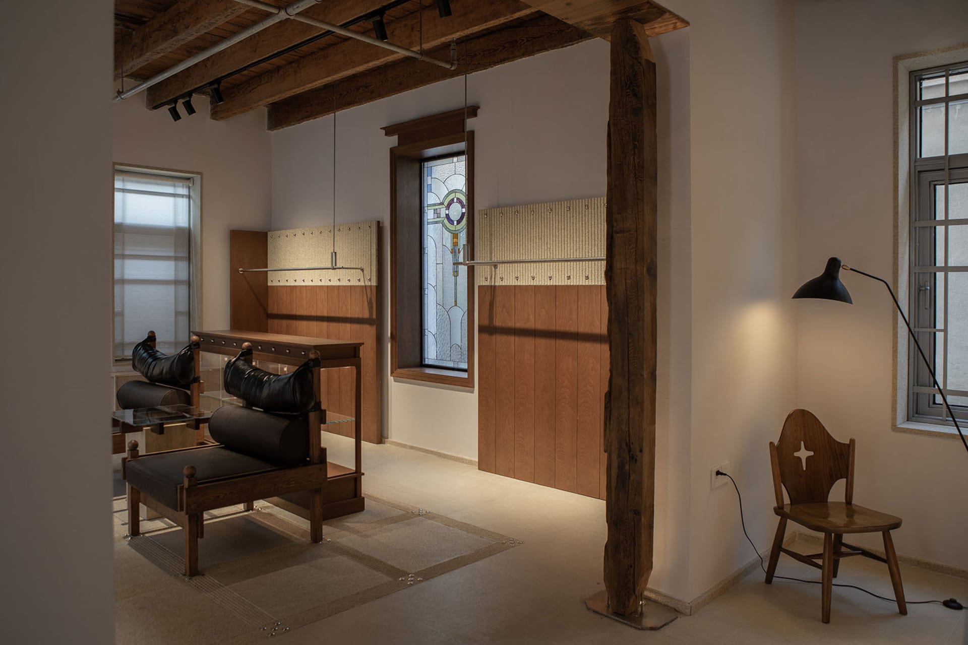 Modern retail interior showcasing large reclaimed wood structural beams, dark leather upholstered benches on glass and wood display tables, wood-paneled walls, and a wooden chair with cross cutout under warm track lighting.