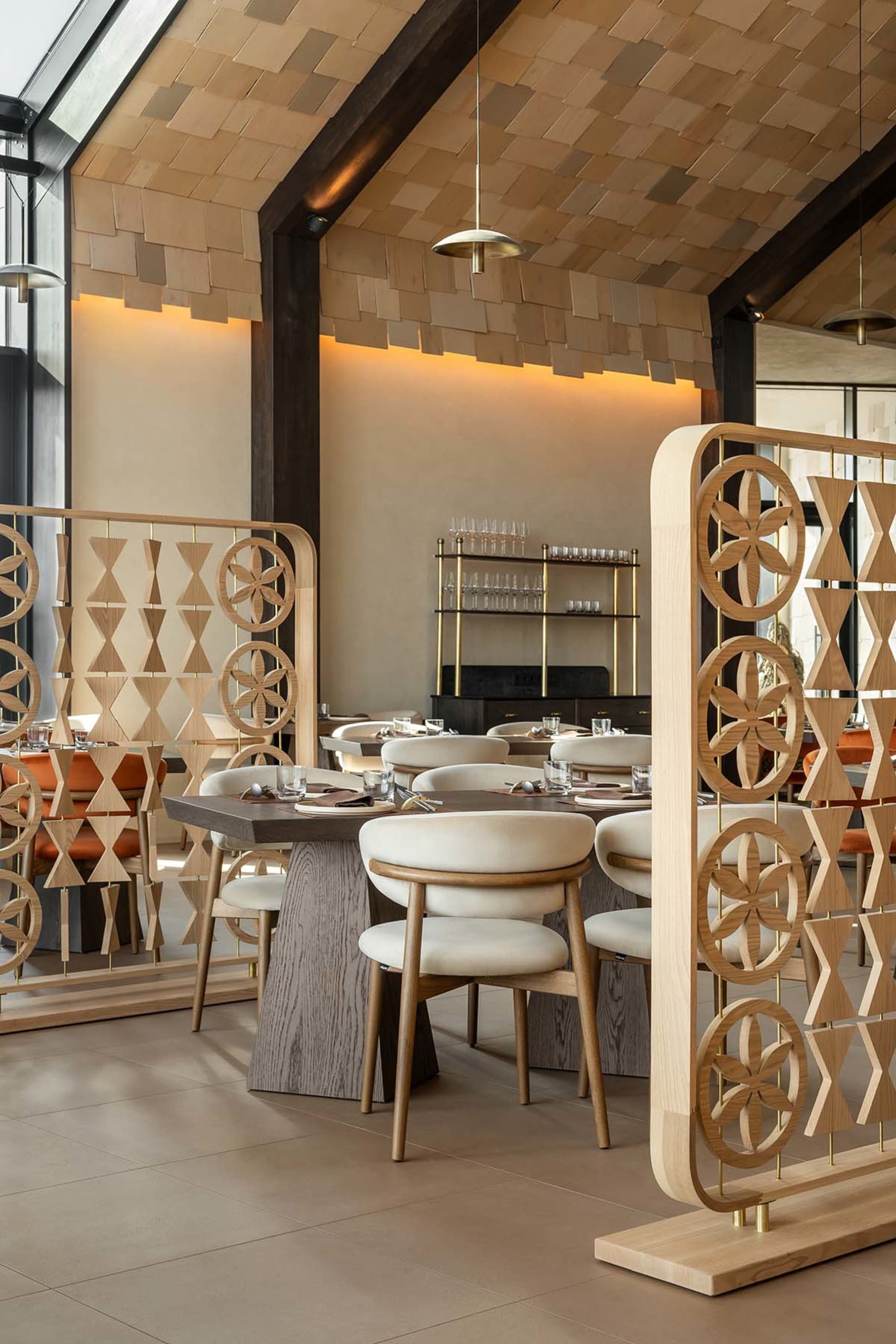 Modern Japanese-inspired restaurant interior featuring light wood dining tables, cream upholstered chairs, and ornate wooden room dividers with floral and geometric patterns under a wooden shingle ceiling.
