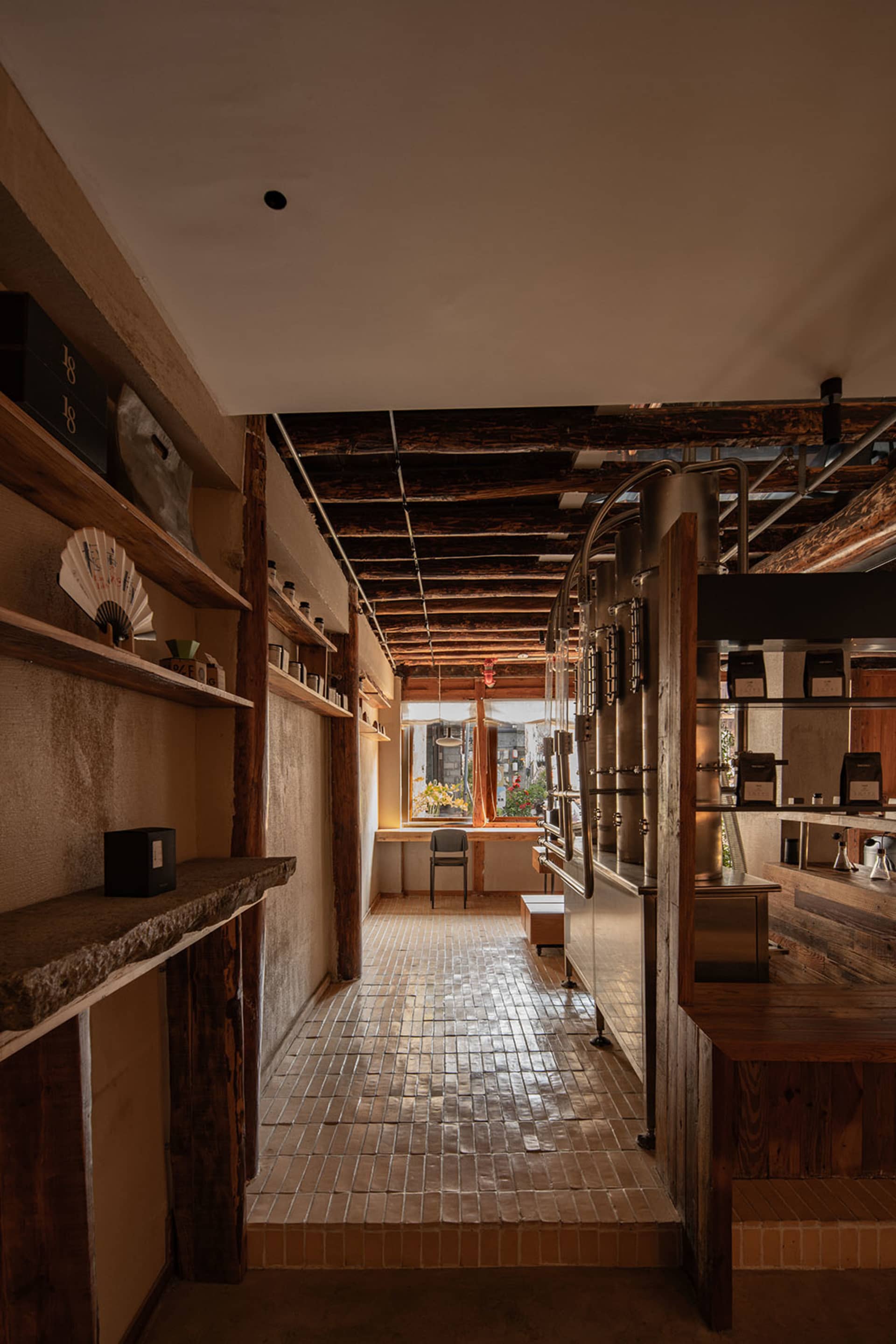Narrow interior corridor of an industrial-style coffee roastery featuring reclaimed wood shelves, exposed dark wooden ceiling beams, terracotta brick floor, and stainless steel roasting equipment on the right.