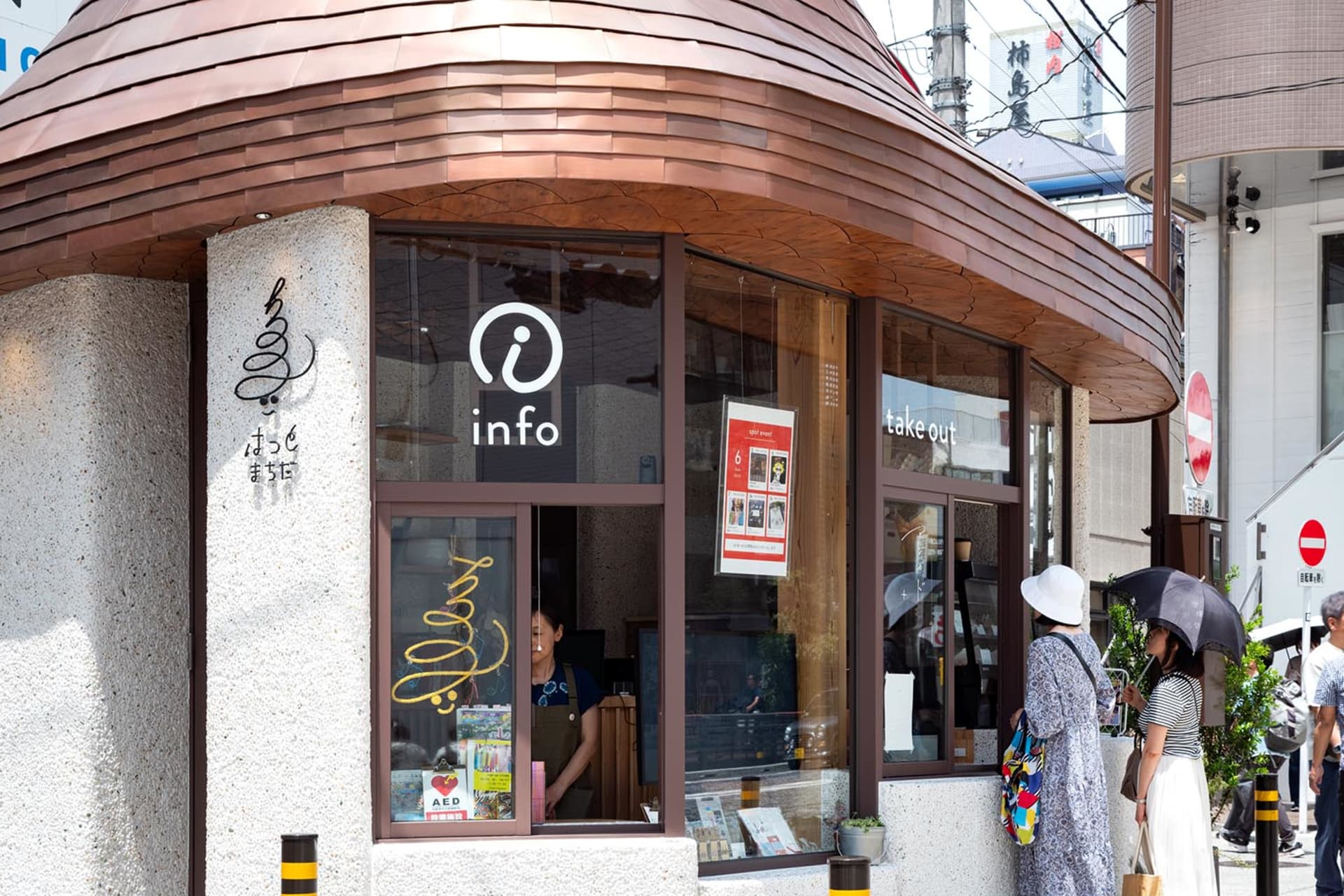 Street view of a Japanese cafe with a distinctive curved copper-tiled roof, textured concrete facade, large glass windows, and customers standing outside on a sunny day.
