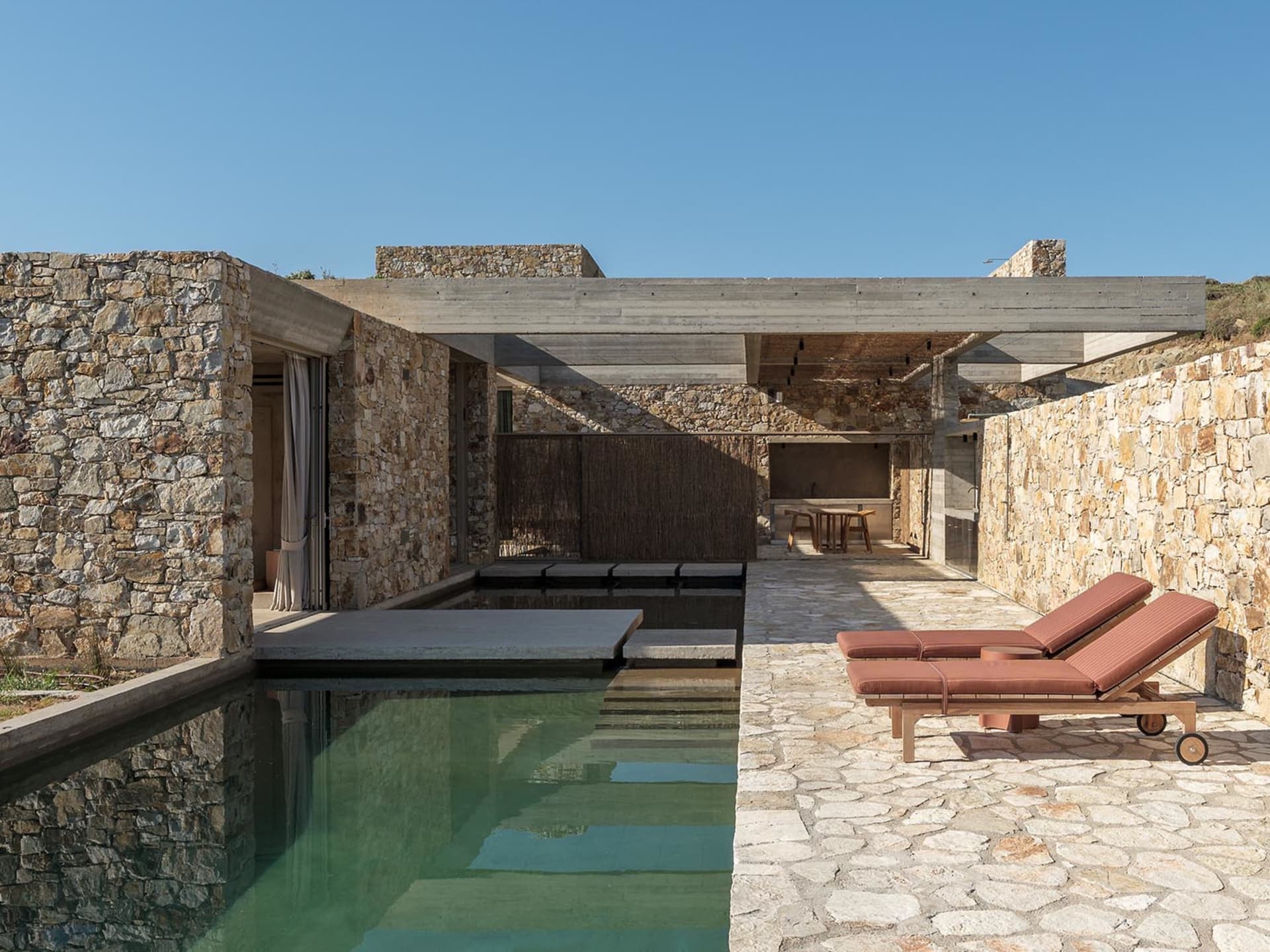 Outdoor stone courtyard featuring a narrow reflecting pool, concrete overhang, two terracotta lounge chairs on a cobblestone patio, and a dining area in the background under a clear blue sky.