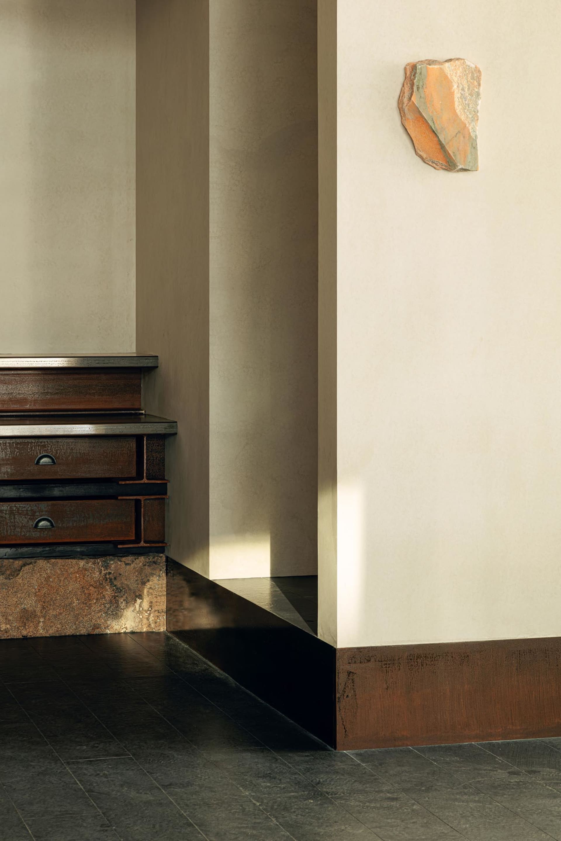 Modern minimalist interior corner featuring a dark corten steel staircase base, dark tiled floor, neutral plaster walls, and an abstract orange stone sculpture mounted on the wall.