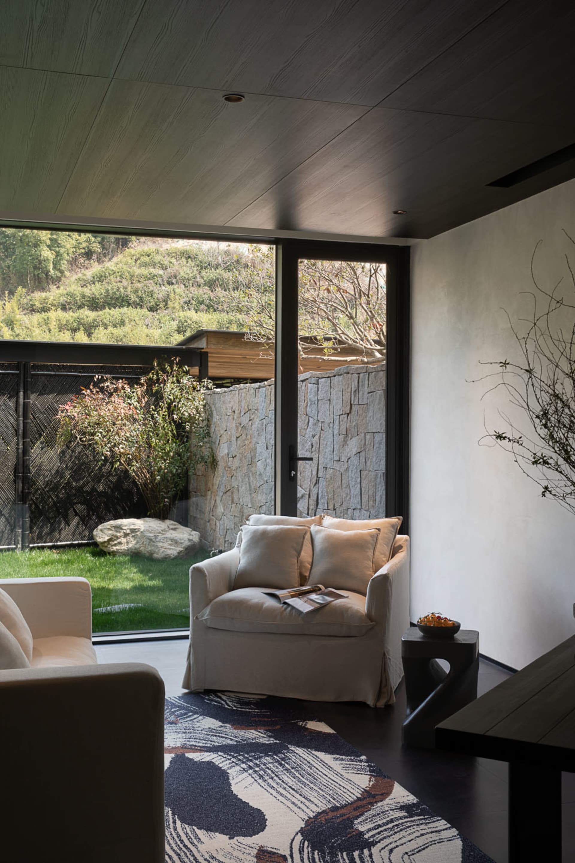 Modern living room interior featuring a cream armchair, dark wood ceiling, stacked stone wall, and large glass doors opening to a green garden with a patterned area rug in the foreground.