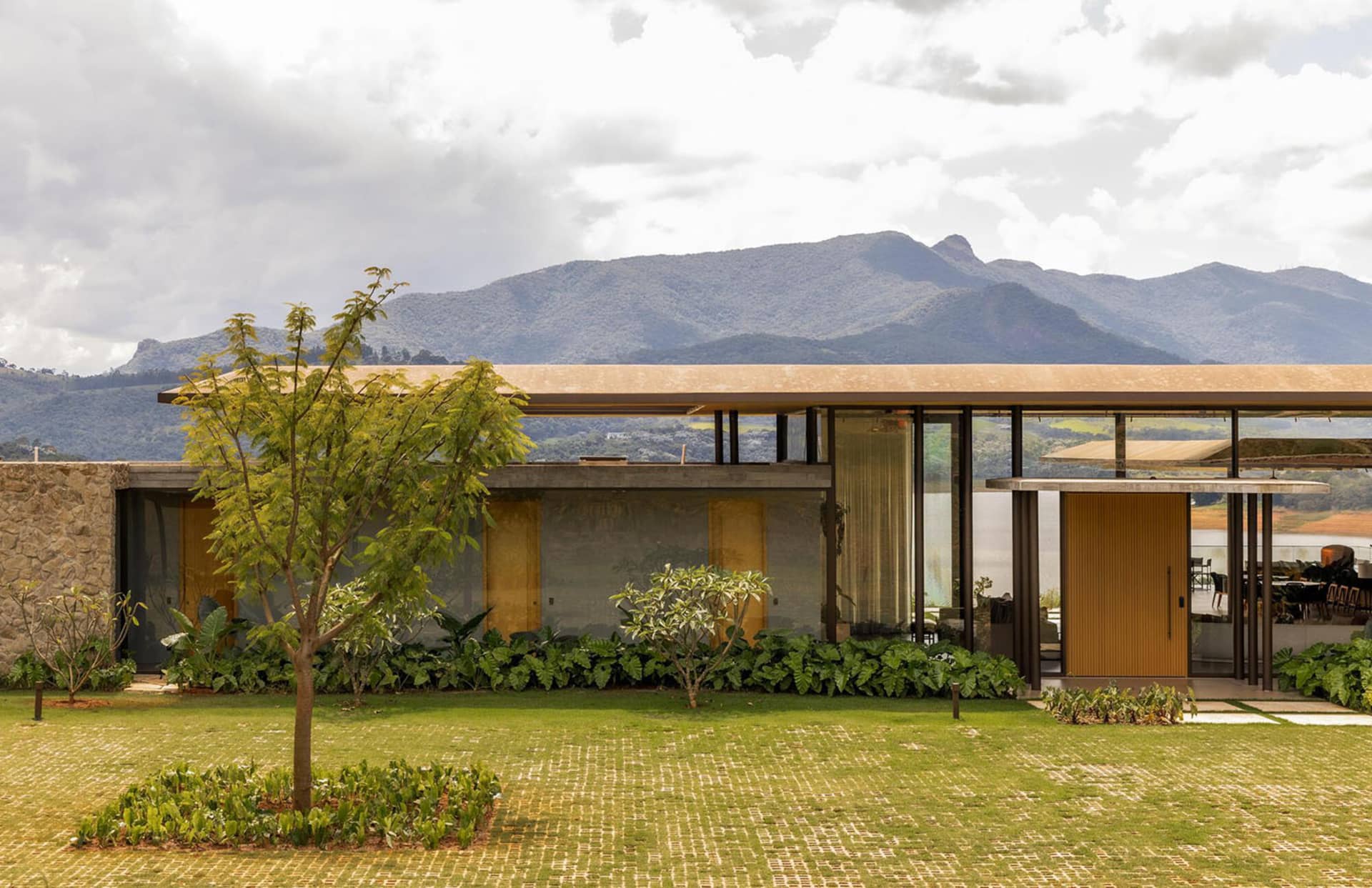 Modern single-story mountain house featuring concrete walls, vertical wood slats, large glass windows, and lush tropical landscaping with a dramatic mountain range in the background.