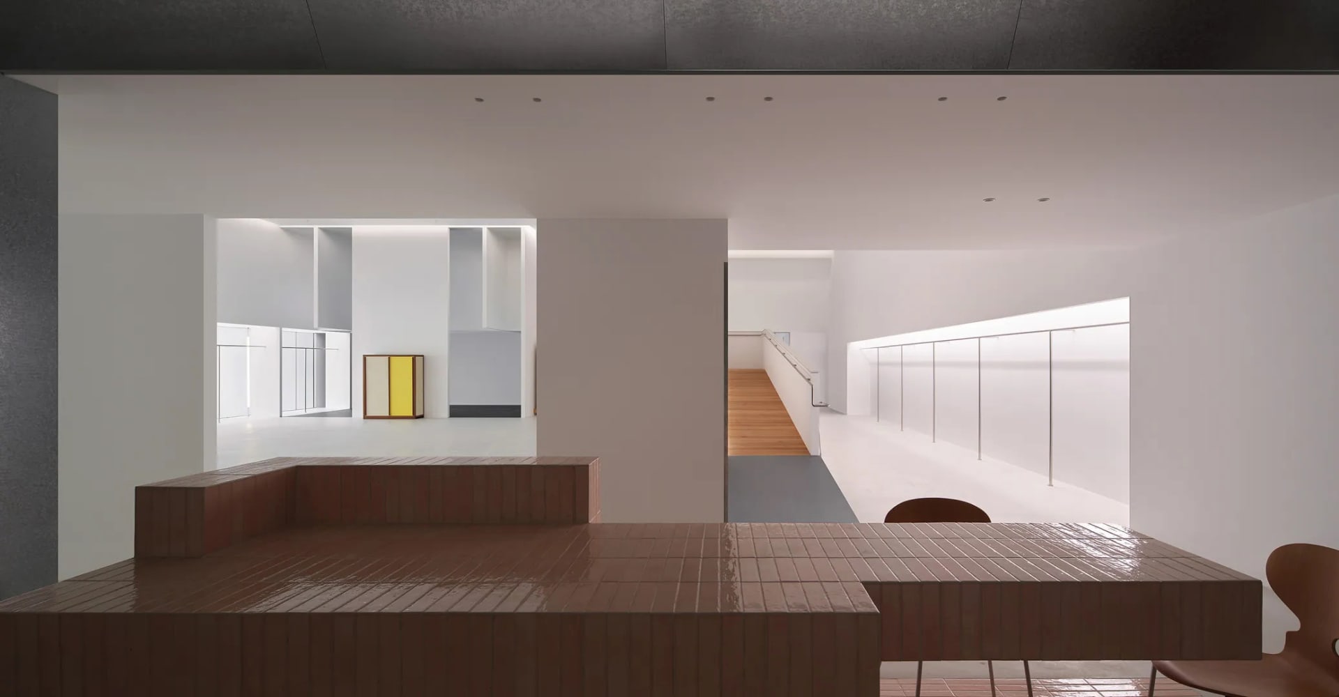 Modern minimalist dining area featuring a large terracotta tiled counter with integrated banquette seating, wooden chairs, white walls, and a view into a long corridor with wooden flooring.