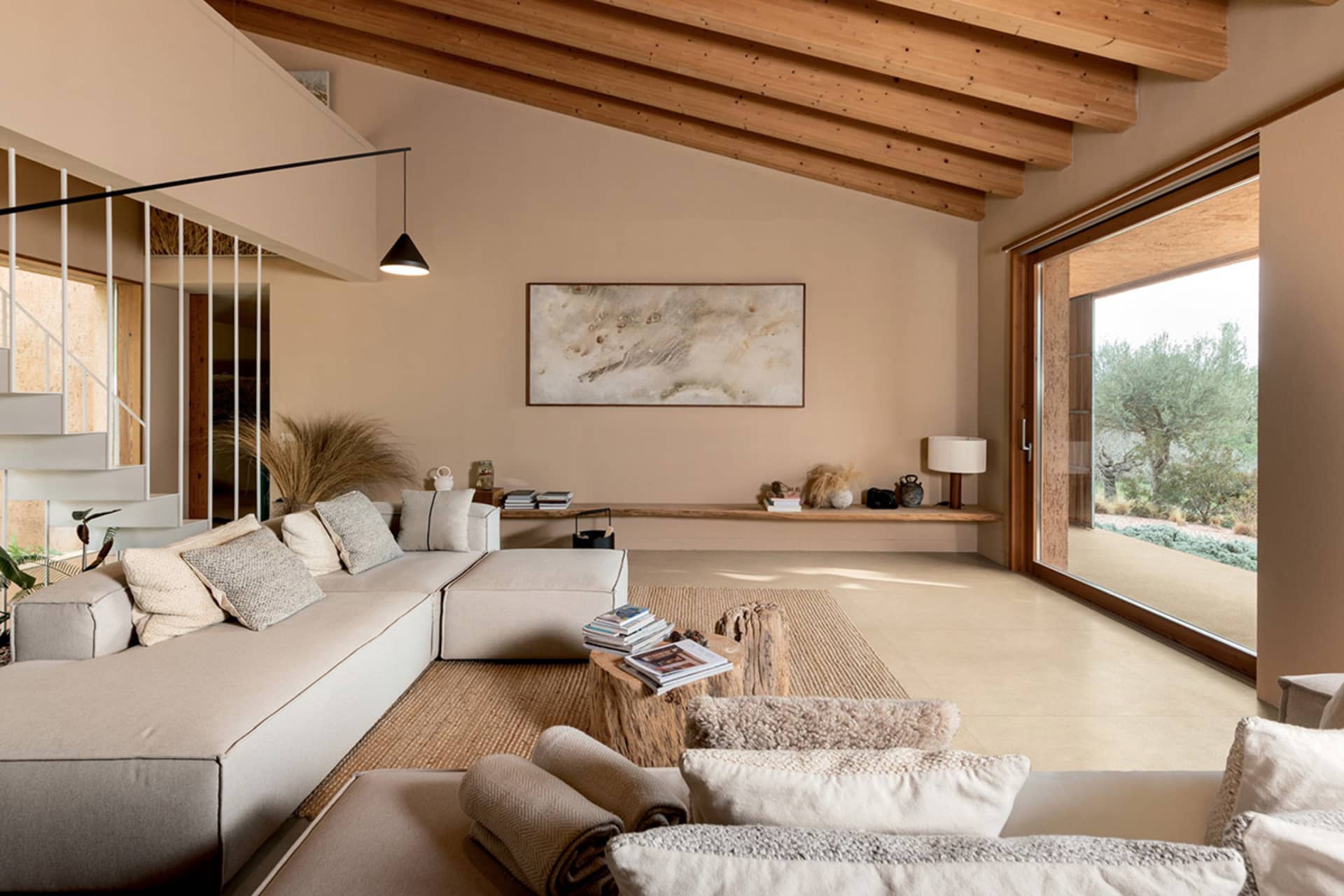 Modern organic living room featuring a large L-shaped neutral sofa, tree-trunk coffee table, exposed wooden ceiling beams, and large sliding glass doors opening to a garden.