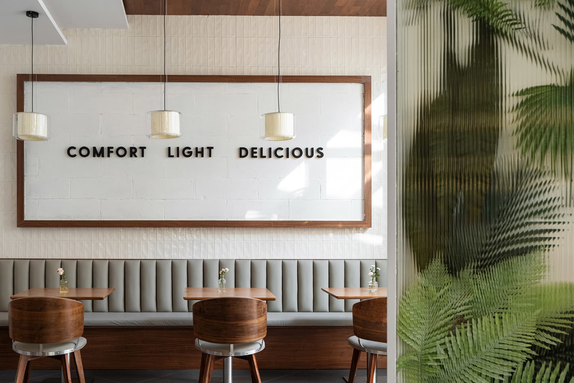 Modern restaurant dining area with grey upholstered booth, round walnut wood chairs, wooden tables, white tiled wall with large wooden frame sign reading 'COMFORT LIGHT DELICIOUS', and three pendant lights.