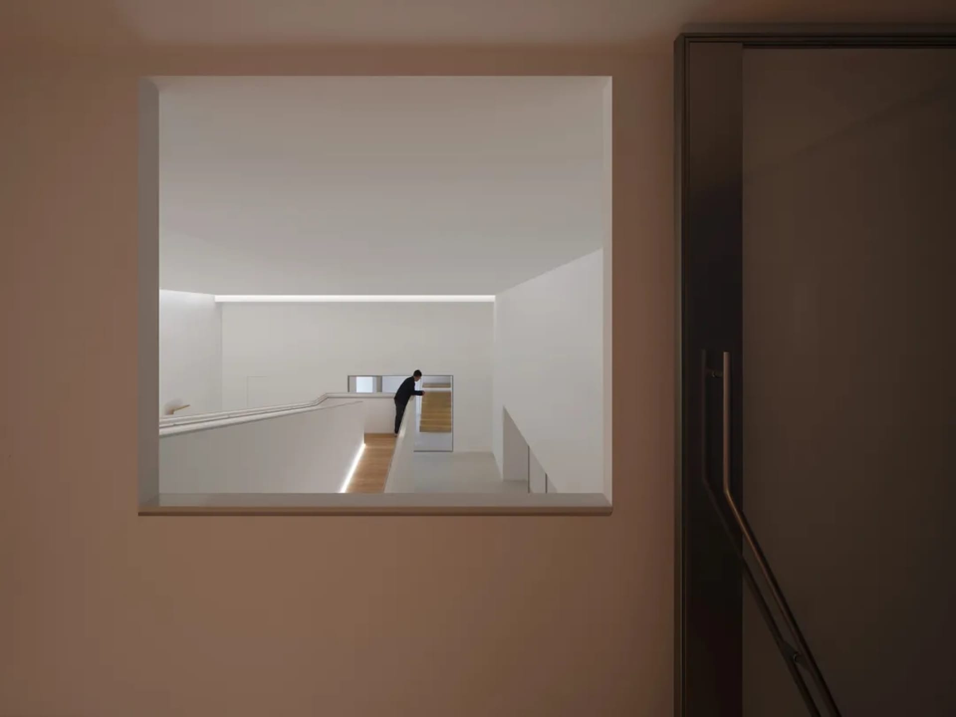 View through a square wall opening into a minimalist white hallway featuring a modern staircase with integrated LED lighting and a person standing at the far end.
