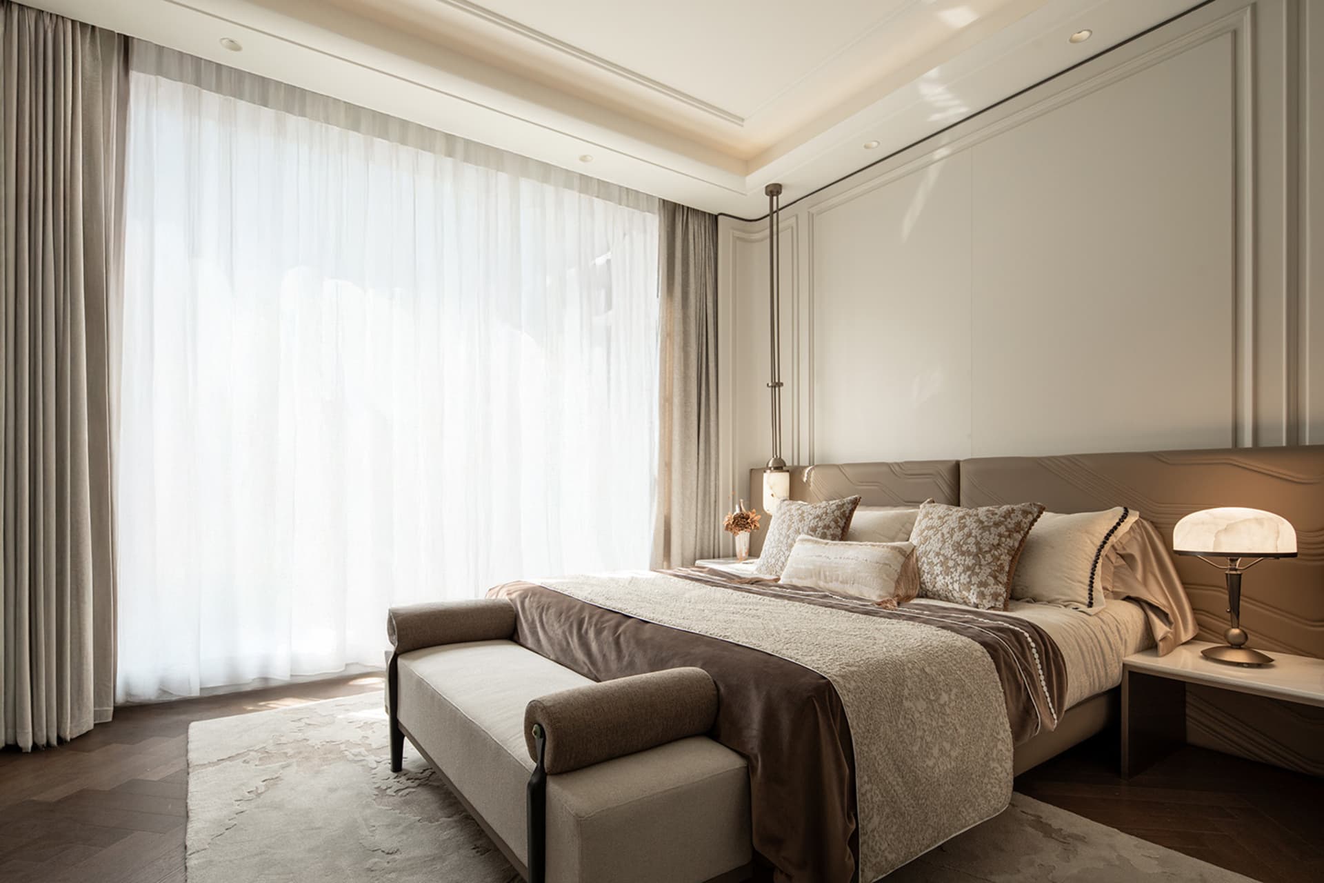 Modern luxury bedroom featuring a sculptural taupe wave headboard, neutral bedding with patterned accent pillows, a pendant light, marble table lamp, and large sheer curtains allowing bright daylight.