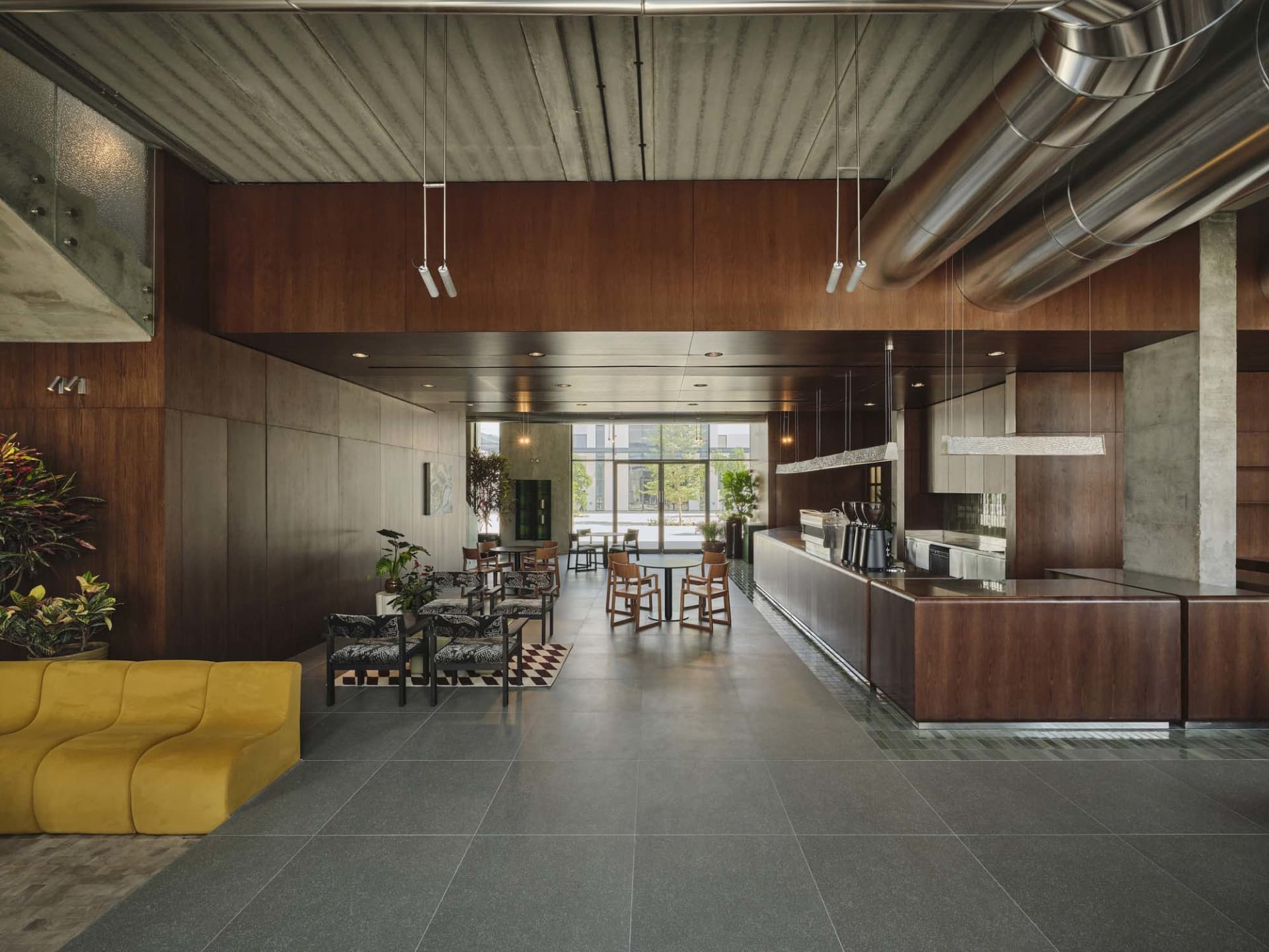 Modern industrial-style commercial cafe lounge with dark walnut wood walls, polished concrete floors, exposed stainless steel ducts, a bright yellow curved sofa in the foreground, patterned armchairs, and a long service counter under large windows.